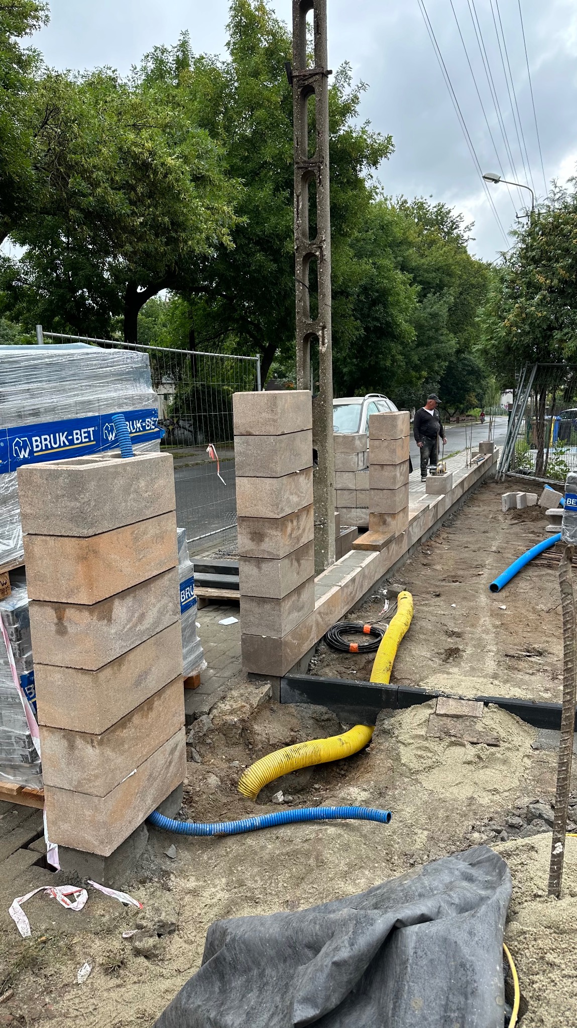 Budowa niskiego murku z betonowych bloczków na zewnątrz, widoczne rury karbowane w kolorze niebieskim i żółtym, paleta z kostką brukową w tle.