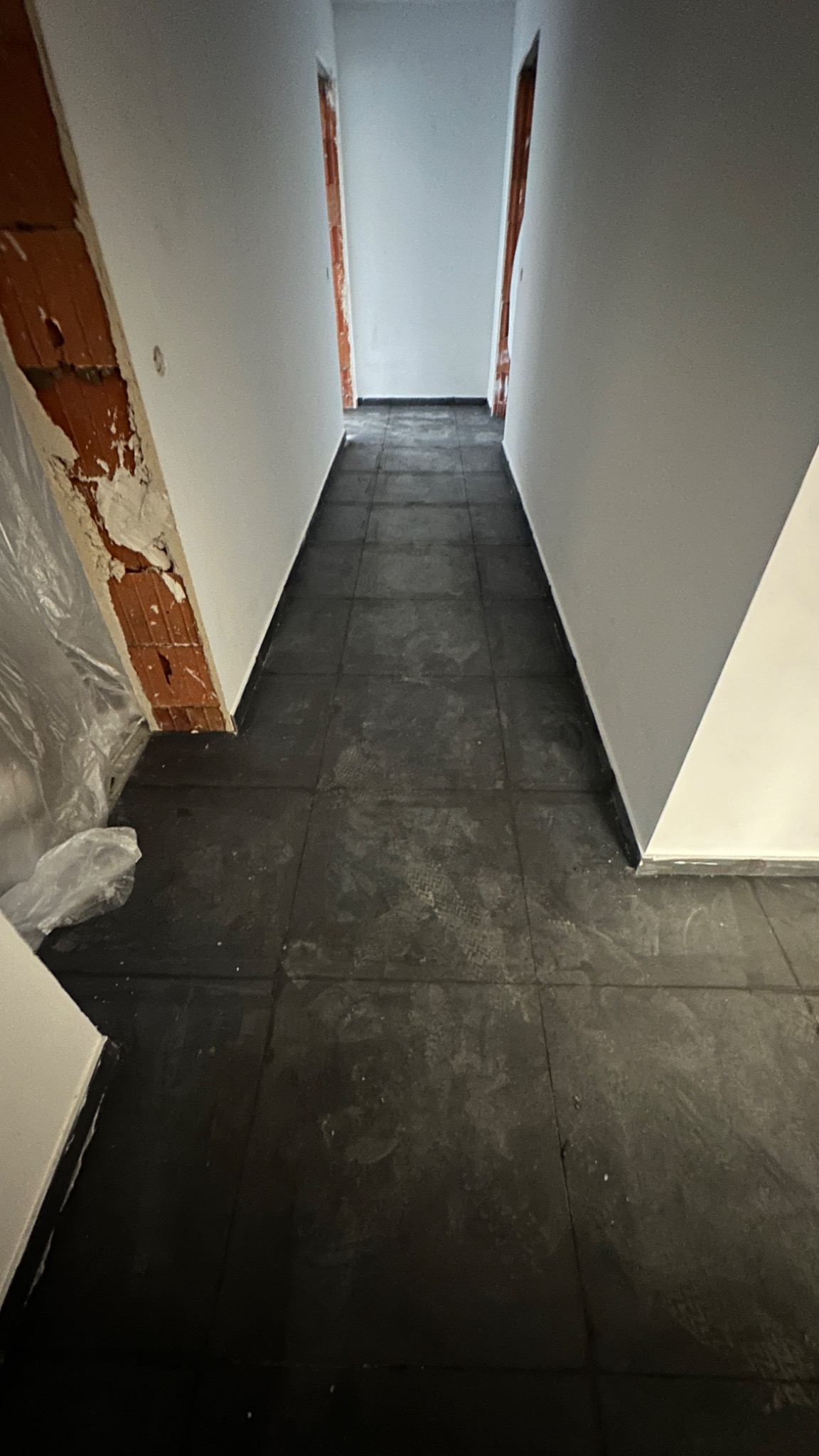 Korytarz w trakcie remontu, widoczne szare płytki podłogowe z zabrudzeniami, surowe ściany w odcieniach bieli i szarości, fragment ściany z odsłoniętą czerwoną cegłą i resztkami tynku zabezpieczony...