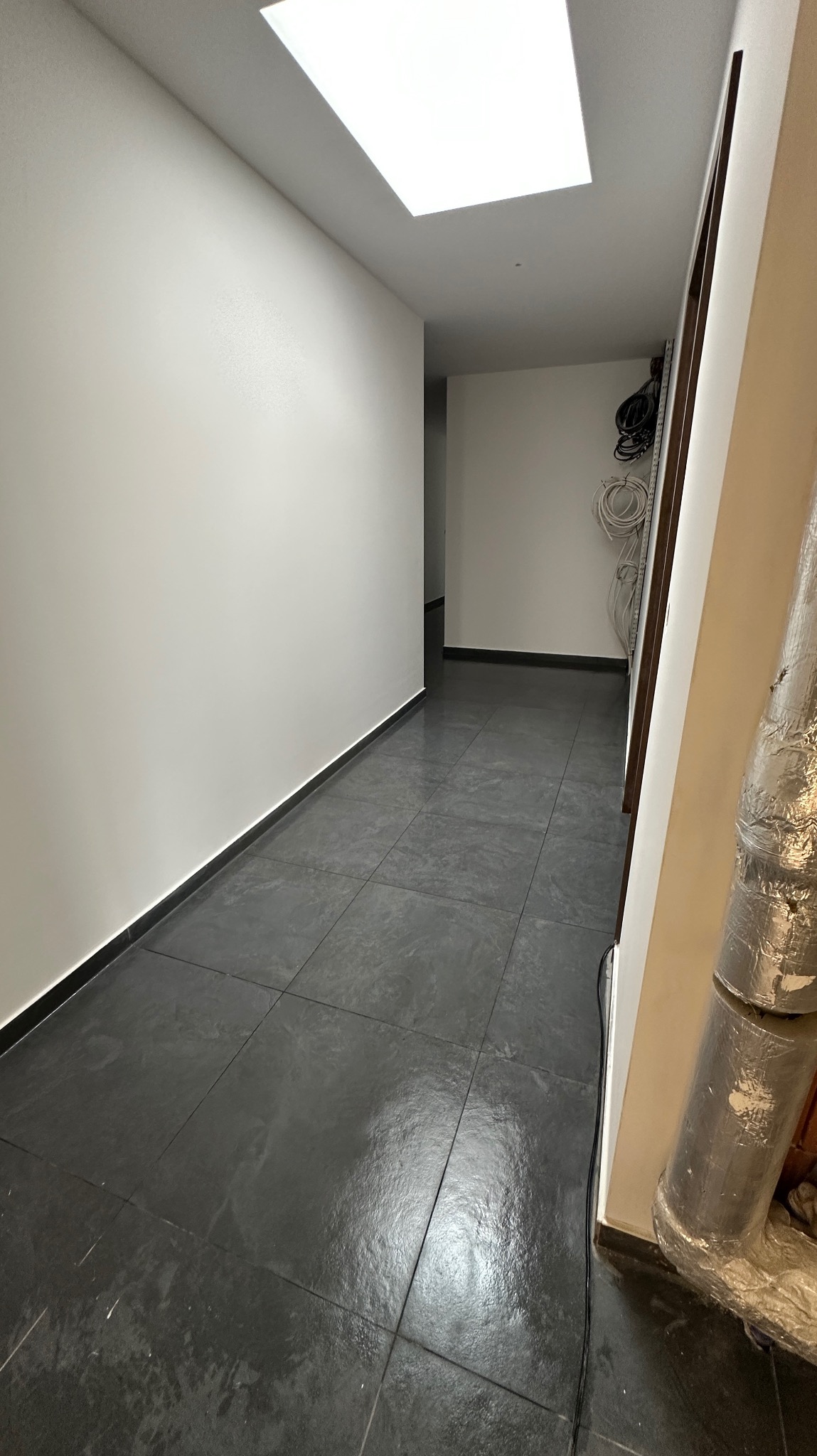 Korytarz z szarymi płytkami podłogowymi, świeżo pomalowane białe ściany, widoczne kable i rury w tle, sufit z wbudowanym oświetleniem.