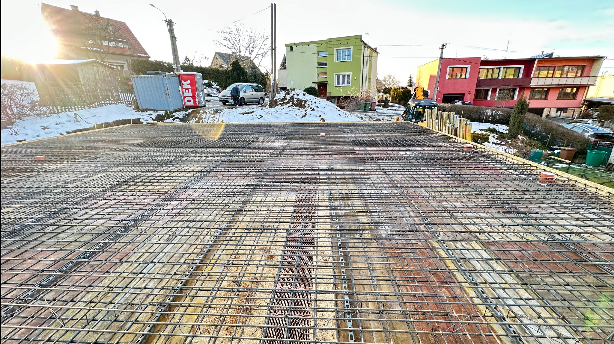 Zbrojenie płyty fundamentowej domu jednorodzinnego, widok z góry na siatkę stalową na deskowaniu, w tle budynki mieszkalne i częściowo odśnieżony teren.