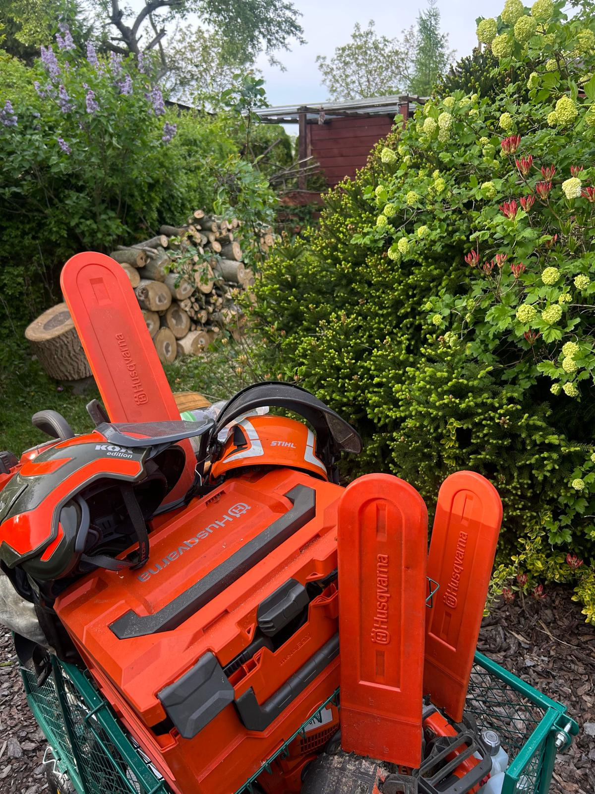 Sprzęt do prac ogrodniczych: pilarki Husqvarna i Stihl, kaski ochronne, skrzynka narzędziowa na tle zieleni i stosu pociętych pni drzew. Przygotowanie do pracy.