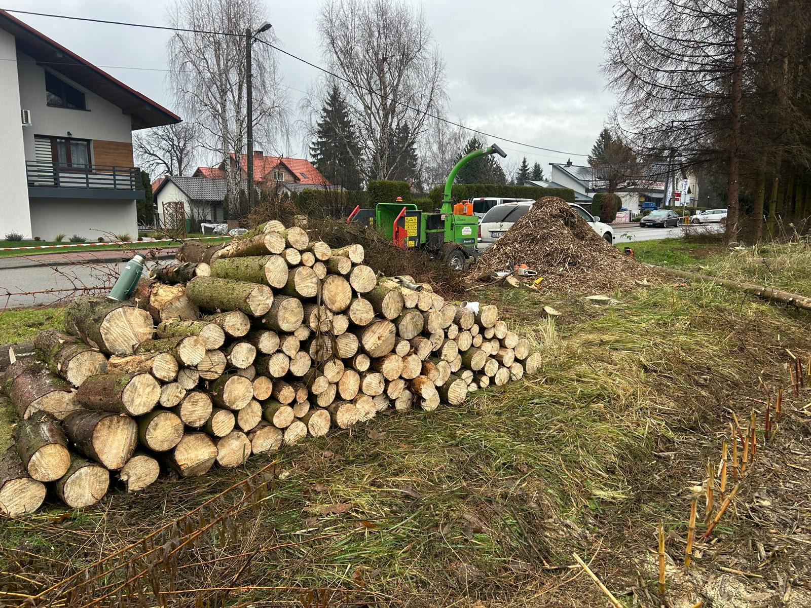 Stos pociętych pni drzew na trawie, w tle rębarka do gałęzi i sterta zrębków. Widoczne budynki mieszkalne i drzewa. Prace porządkowe po usunięciu drzew.