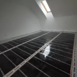 Poddasze z oknem dachowym, na podłodze rozłożona folia grzewcza marki Cronos, przygotowana do dalszych prac wykończeniowych.