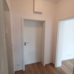 Budomeb - Białe drzwi wewnętrzne z chromowaną klamką w świeżo odnowionym korytarzu z panelami podłogowymi i białymi ścianami, widoczny fragment wentylacji pod sufitem.