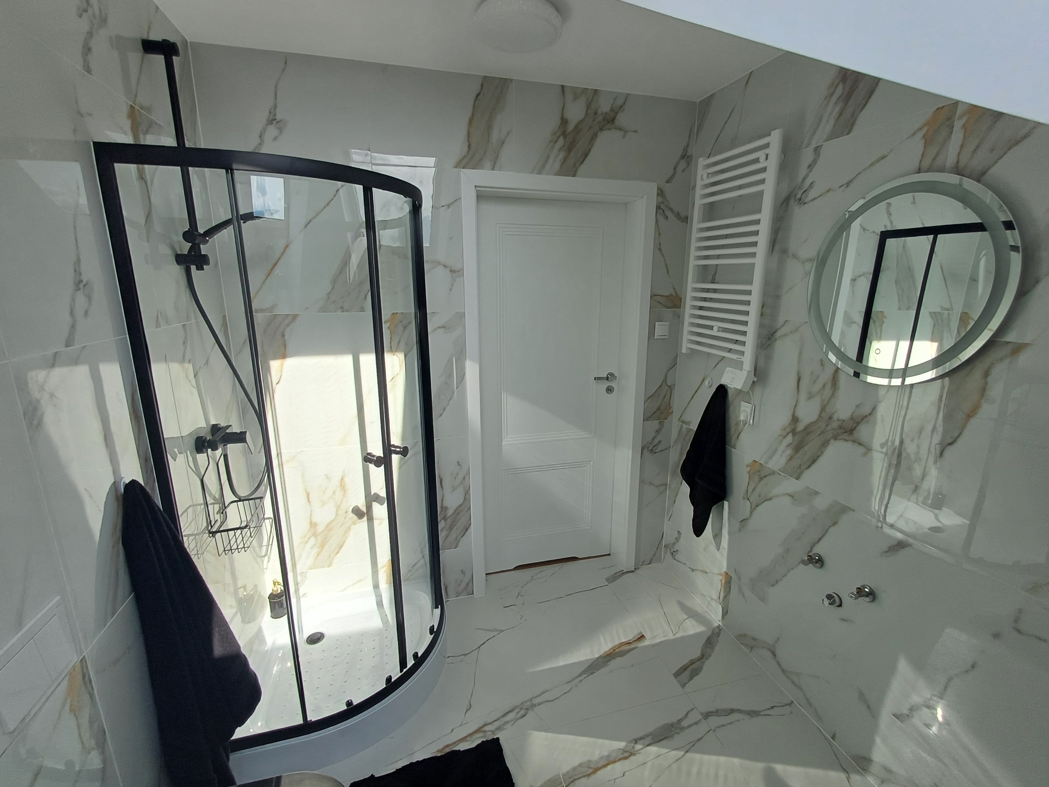 Nowoczesna łazienka z kabiną prysznicową w czarnej ramie, marmurowe płytki na ścianach i podłodze, okrągłe lustro z podświetleniem LED, biały grzejnik drabinkowy i białe drzwi.