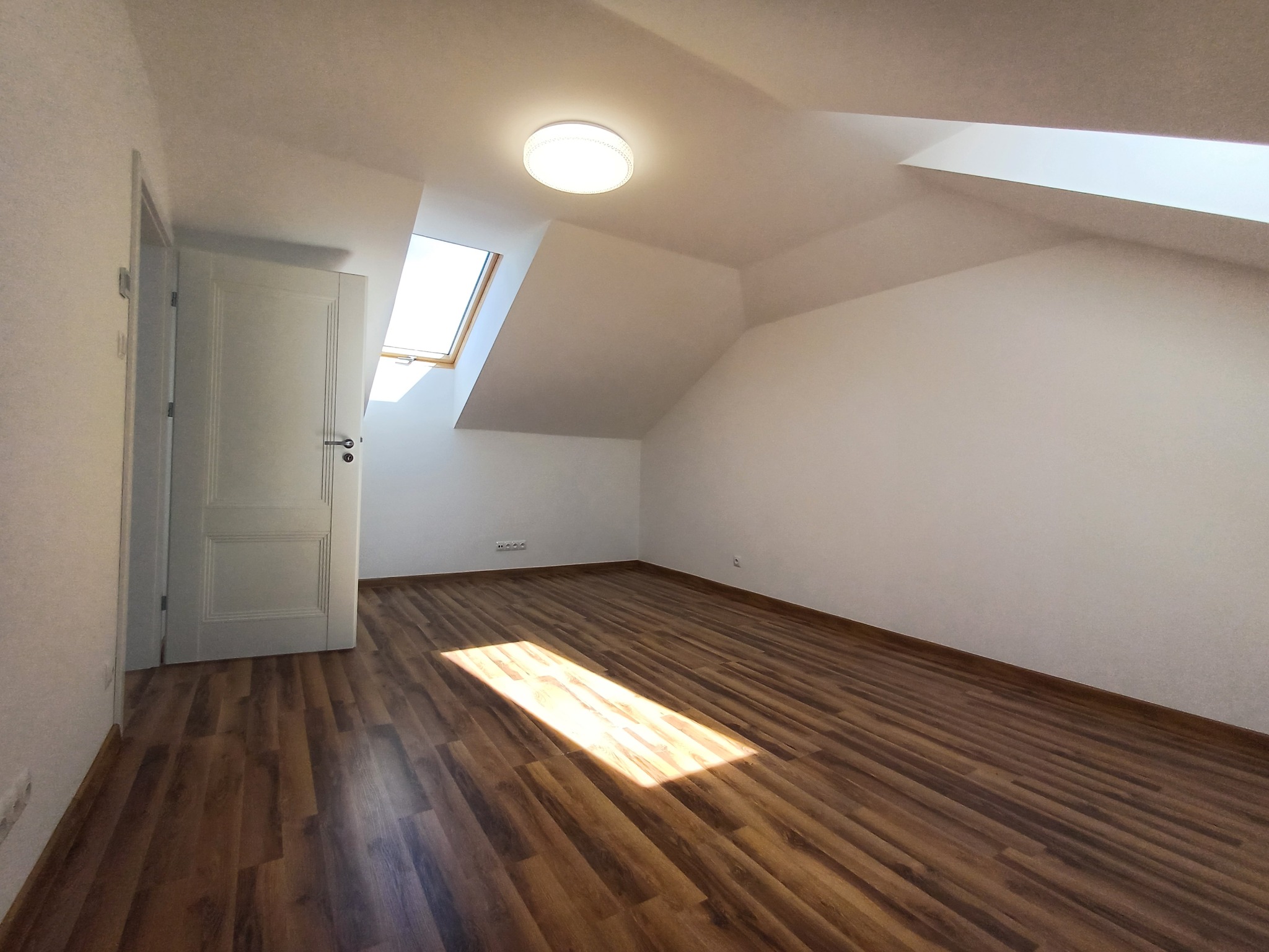 Puste, jasne poddasze z brązowymi panelami podłogowymi, białymi ścianami, oknem dachowym i otwartymi białymi drzwiami.