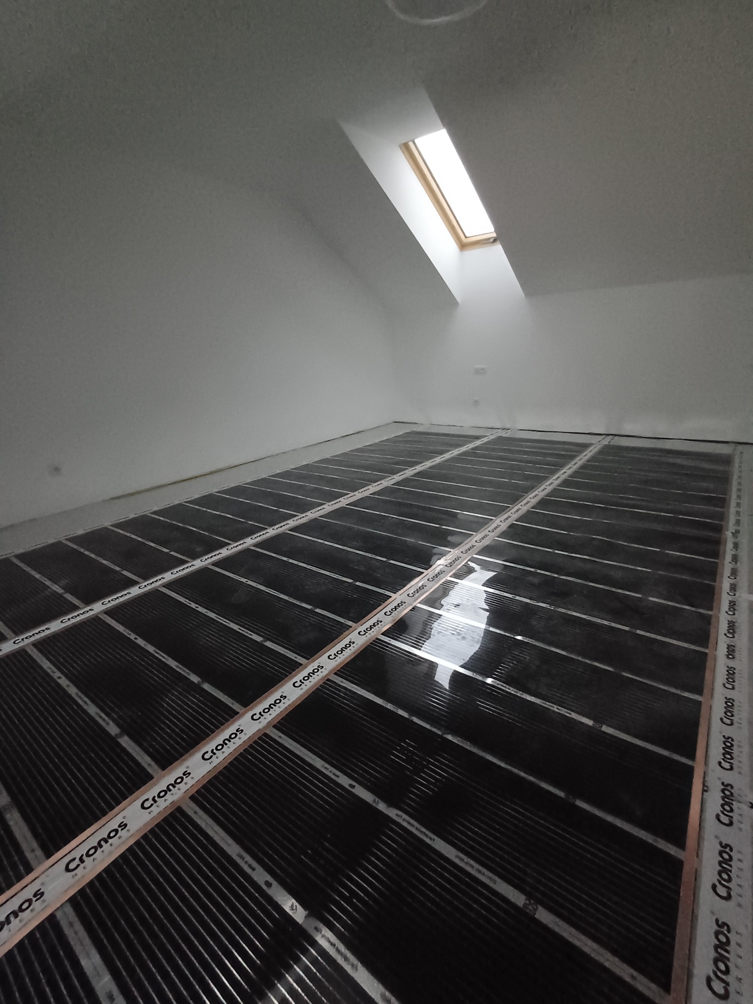 Poddasze z oknem dachowym, na podłodze rozłożona folia grzewcza marki Cronos, przygotowana do dalszych prac wykończeniowych.