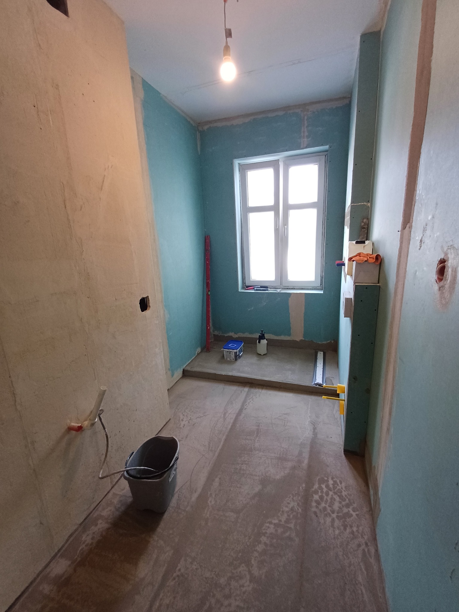 Łazienka w trakcie prac przygotowawczych: ściany częściowo pokryte gładzią, widoczne narzędzia, poziomica oparta o ścianę, wiadro na podłodze.