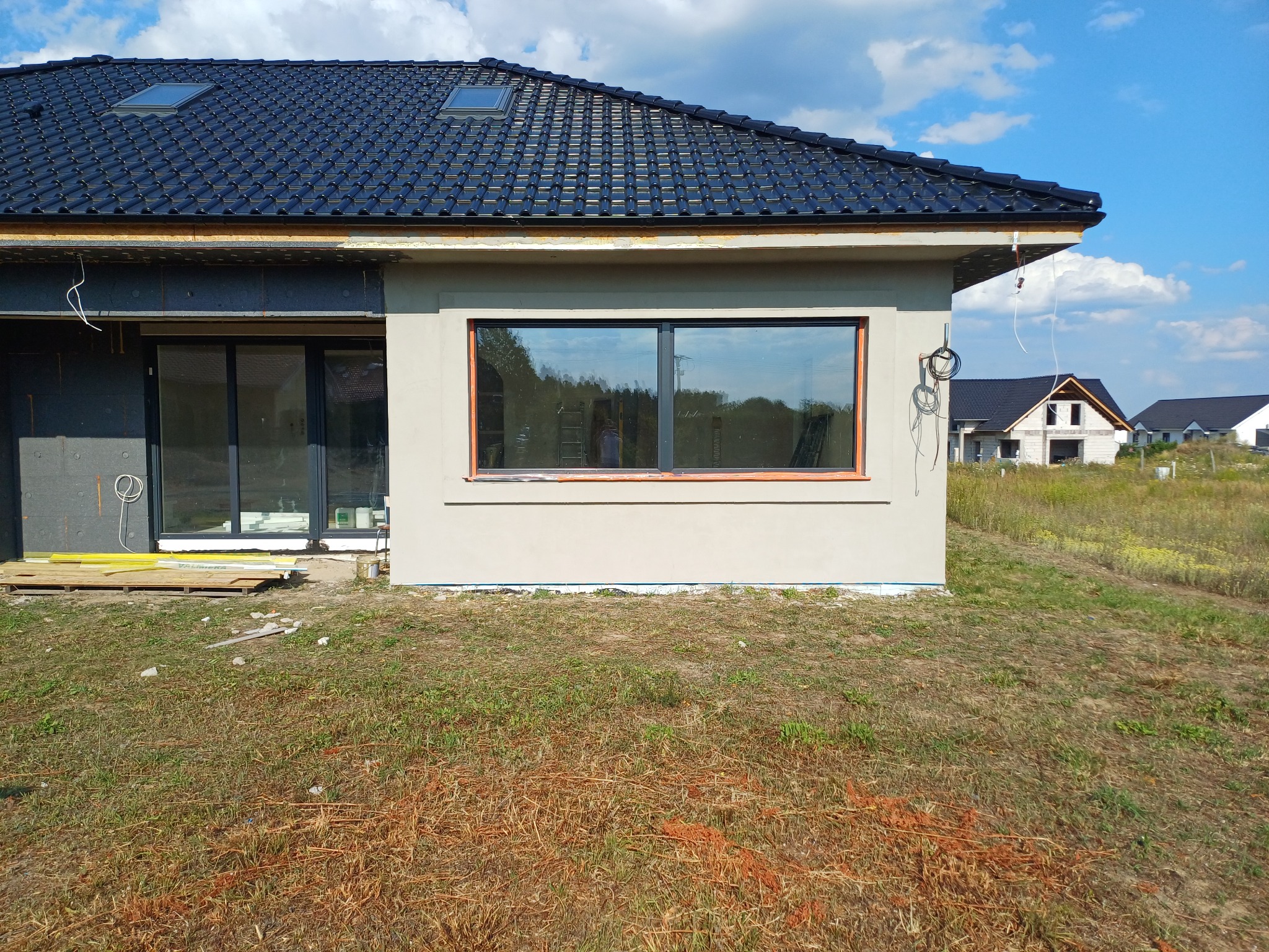 Fragment elewacji domu jednorodzinnego z oknem, po szpachlowaniu, z widoczną izolacją termiczną na ścianie obok, w tle inne budynki w stanie surowym.