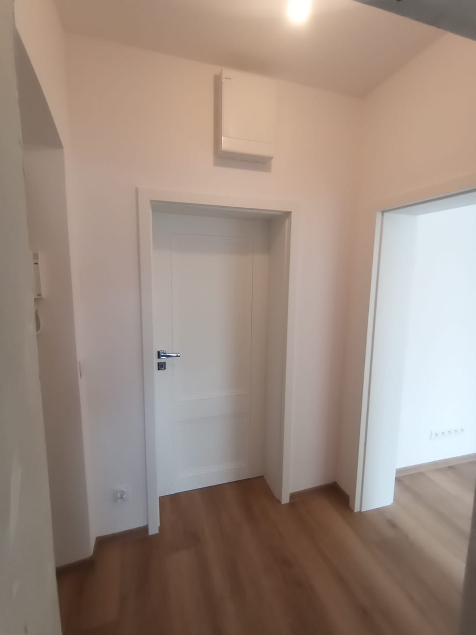 Białe drzwi wewnętrzne z chromowaną klamką w świeżo odnowionym korytarzu z panelami podłogowymi i białymi ścianami, widoczny fragment wentylacji pod sufitem.