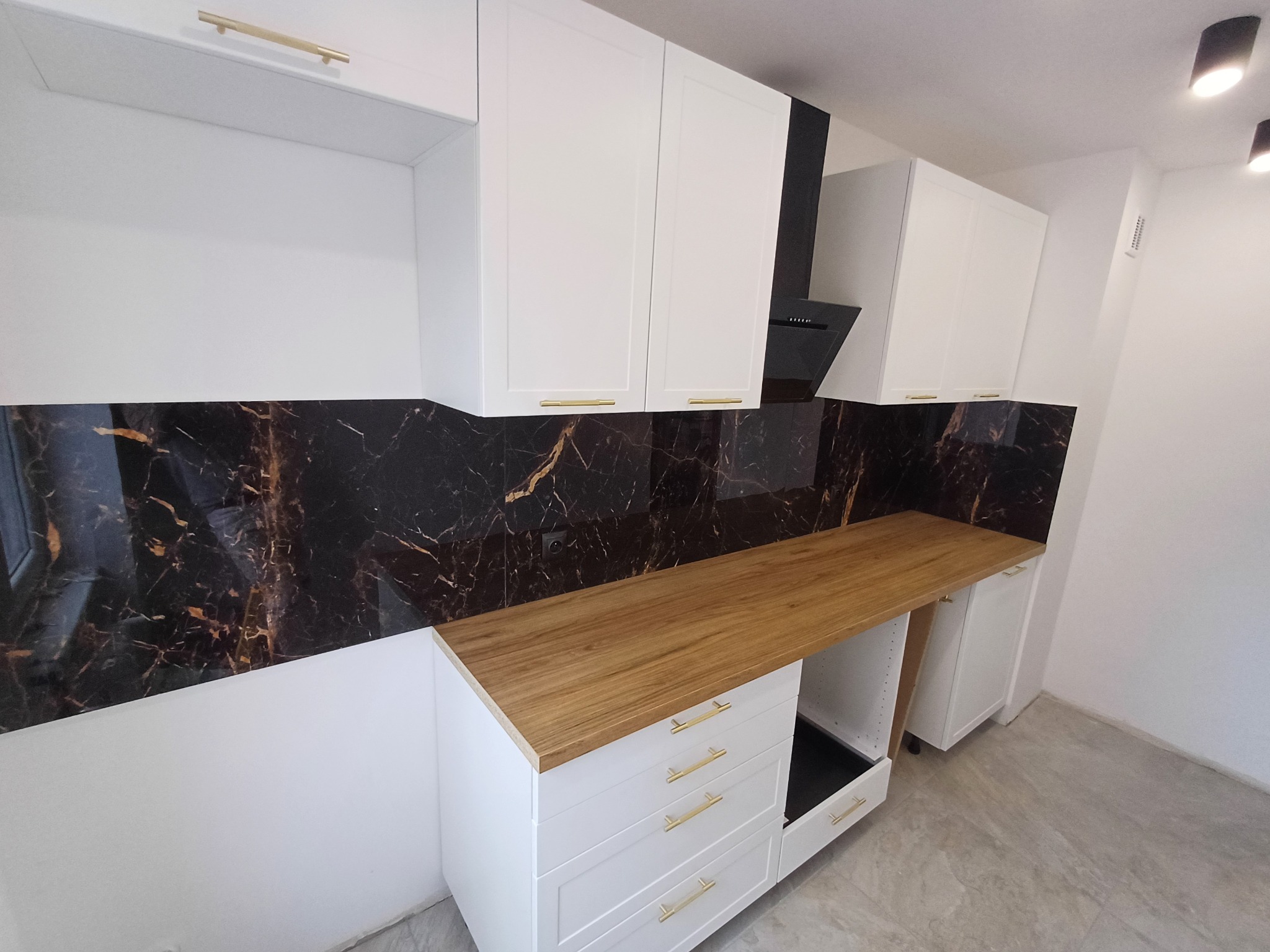 Nowoczesna kuchnia z białymi szafkami, złotymi uchwytami, drewnianym blatem i czarnym panelem ściennym imitującym marmur, widziana pod kątem.