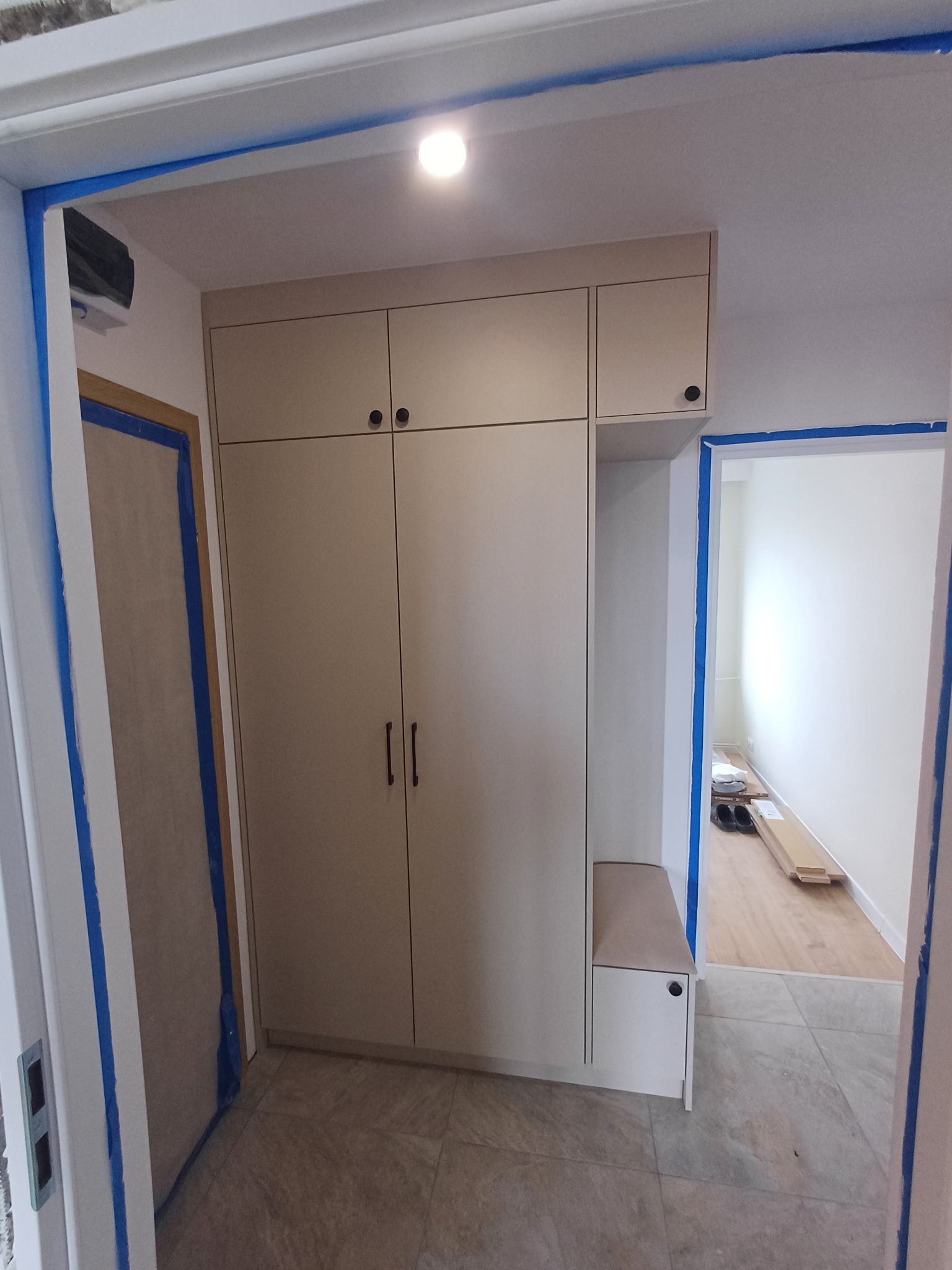 Beżowa szafa wnękowa z czarnymi uchwytami i siedziskiem, zabezpieczona niebieską taśmą malarską na ścianach i ościeżnicach w trakcie remontu mieszkania.