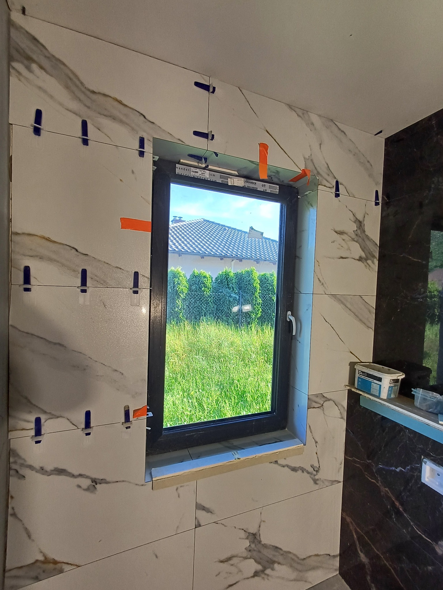 Układanie płytek imitujących marmur wokół okna w łazience, widoczne klipsy poziomujące, za oknem trawnik i dom.