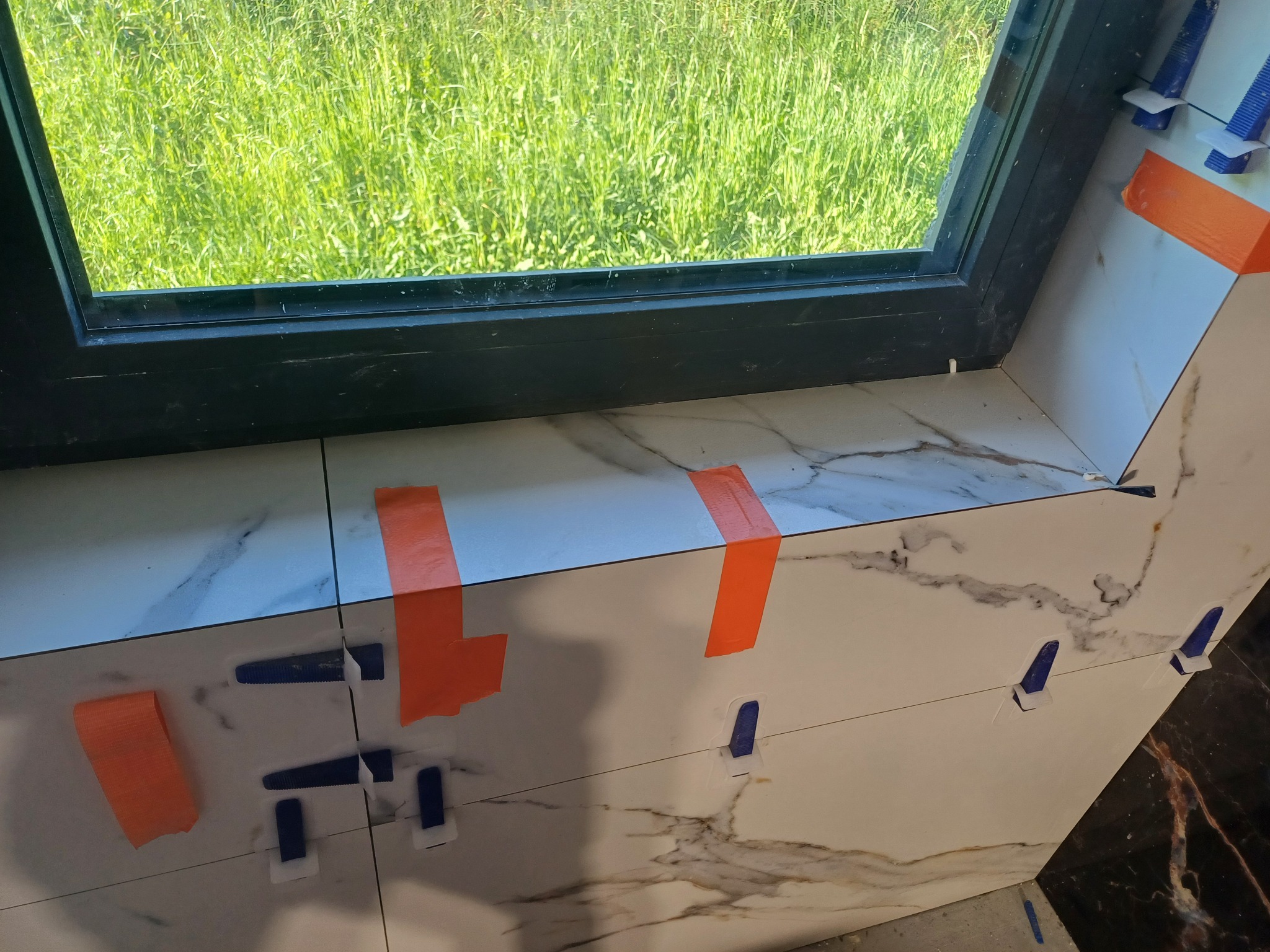 Montaż marmurowych płytek przy oknie, zabezpieczonych pomarańczową taśmą i niebieskimi krzyżykami dystansowymi.