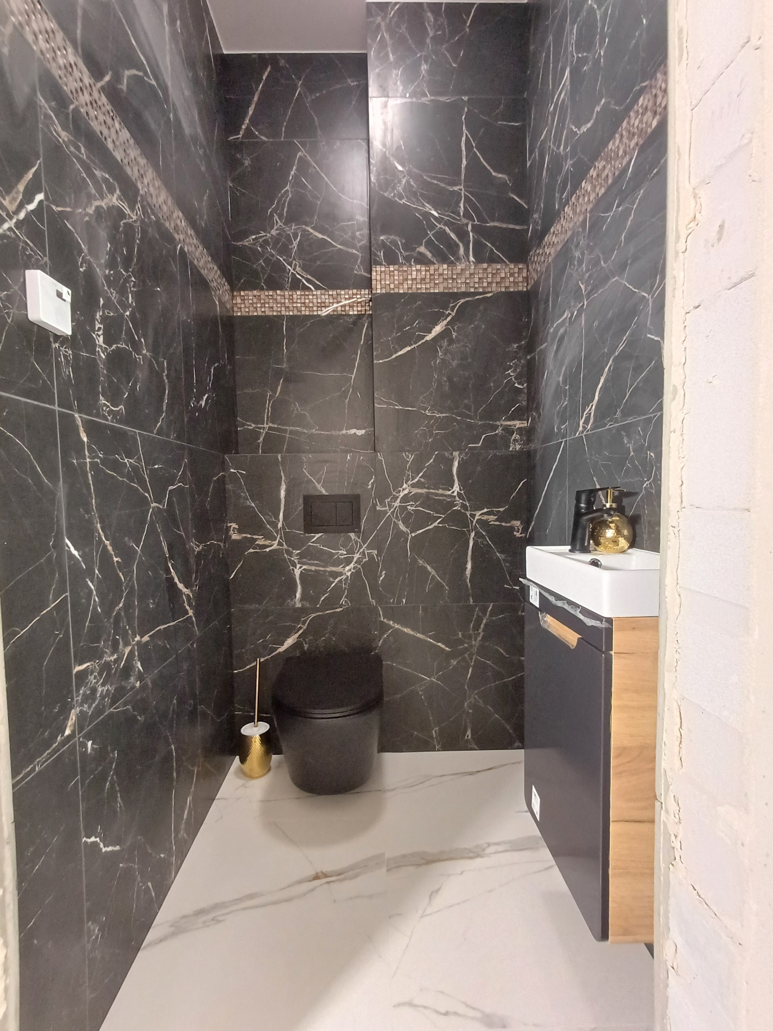 Elegancka łazienka w czarno-białej kolorystyce z marmurowymi płytkami na ścianach, czarną toaletą i umywalką z szafką w odcieniach szarości i drewna, złote akcenty dekoracyjne.