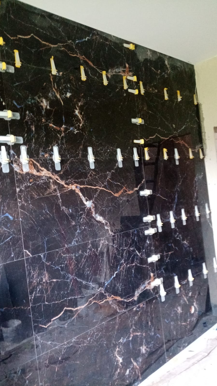 Ściana wyłożona czarnymi płytkami imitującymi marmur z widocznymi klipsami i klinami poziomującymi podczas montażu.