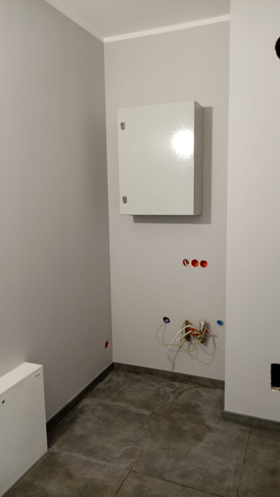 Szara łazienka w trakcie remontu: widoczna biała skrzynka elektryczna na ścianie, przygotowane otwory na gniazdka i instalację wodną, oraz szare płytki podłogowe.