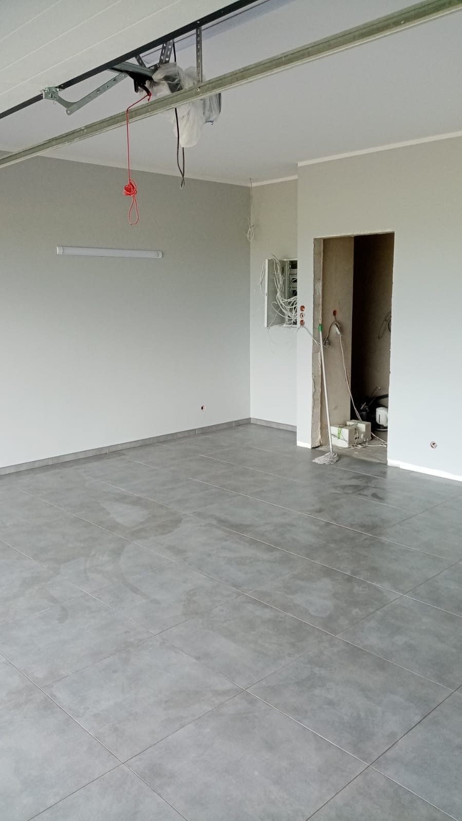 Puste pomieszczenie z szarymi płytkami podłogowymi, świeżo pomalowanymi szarymi ścianami, z otwartą puszką elektryczną i mopem przy wejściu do sąsiedniego pokoju, widoczne elementy konstrukcji sufitu.