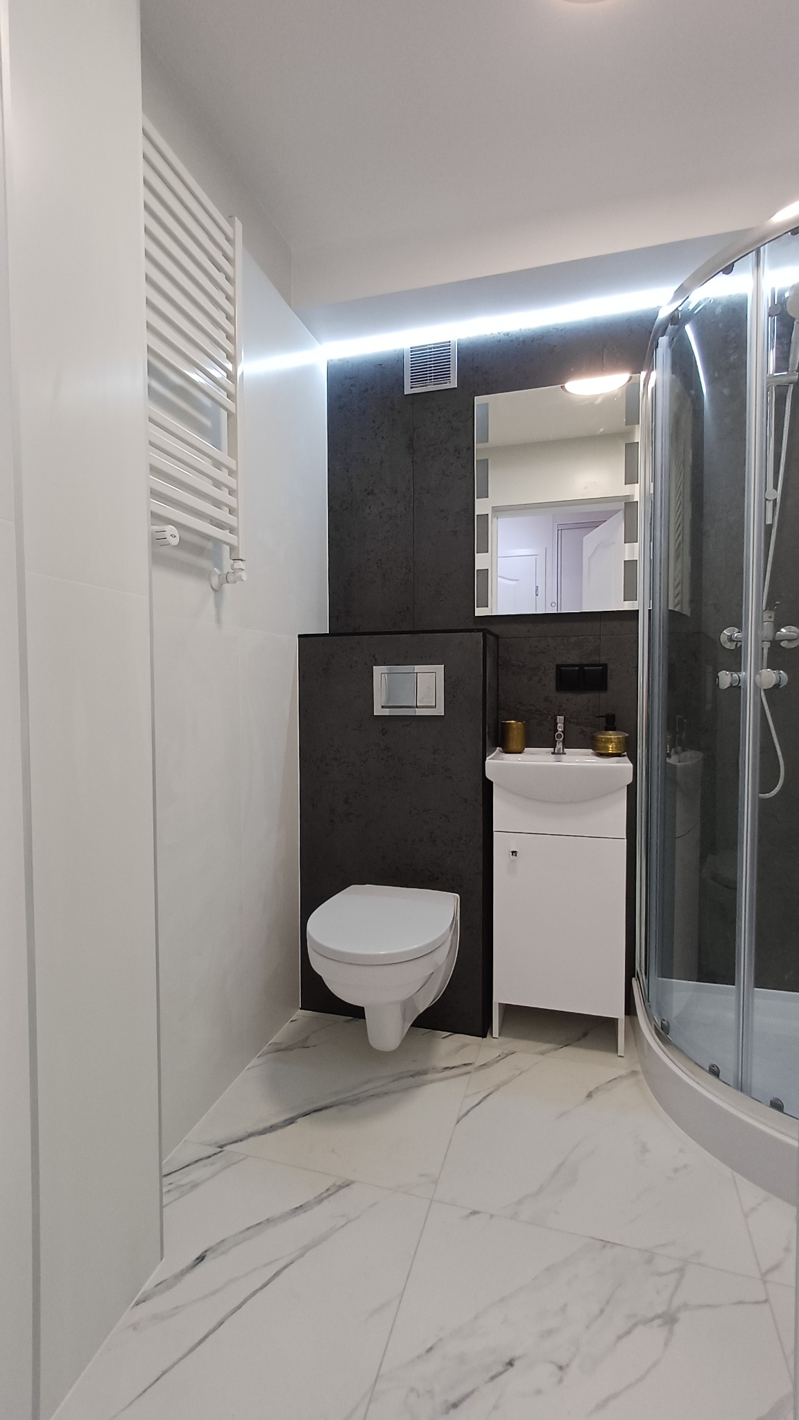 Nowoczesna łazienka po remoncie: biała armatura (toaleta podwieszana, umywalka z szafką), kabina prysznicowa, grzejnik drabinkowy, szare płytki i marmurowa podłoga.