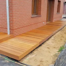 Świeżo wybudowany, gładki taras z jasnego drewna przylegający do ceglanego budynku, widoczne przygotowane podłoże z czarną agrowłókniną i piaskiem.