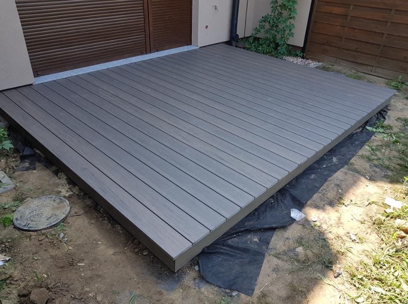 Nowoczesny taras z kompozytowych desek w odcieniach szarości, ułożony bezpośrednio na gruncie z widoczną czarną geowłókniną, obok studzienka kanalizacyjna.