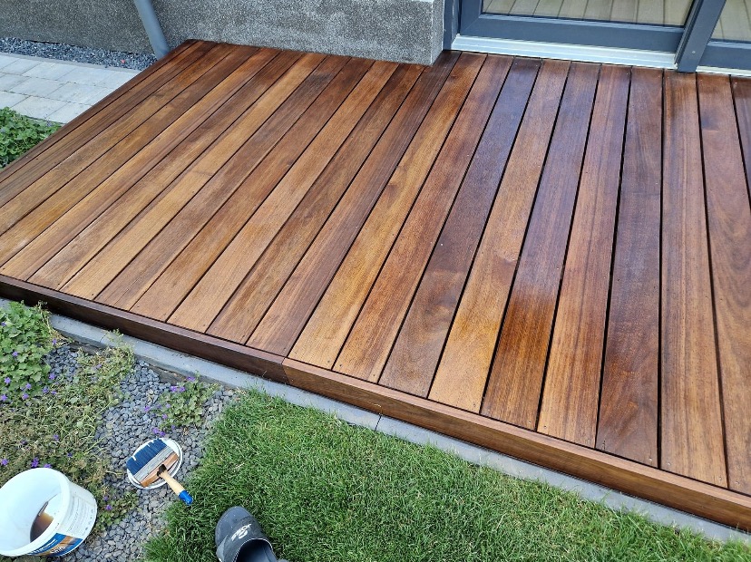 Świeżo polakierowany taras z drewna egzotycznego, widoczny pędzel i wiadro z lakierem, obok trawnik i stopa w klapku.