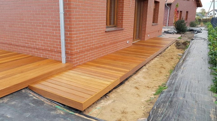 Świeżo wybudowany, gładki taras z jasnego drewna przylegający do ceglanego budynku, widoczne przygotowane podłoże z czarną agrowłókniną i piaskiem.