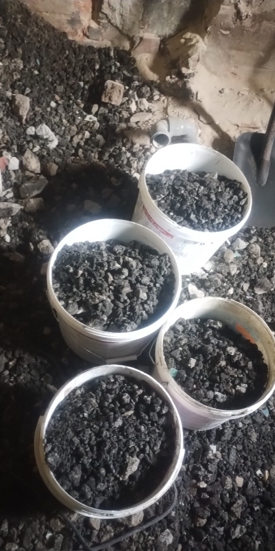 Cztery białe wiadra wypełnione gruzem po wyburzeniach, ustawione na podłodze z ceglaną ścianą w tle i fragmentem rury PCV.