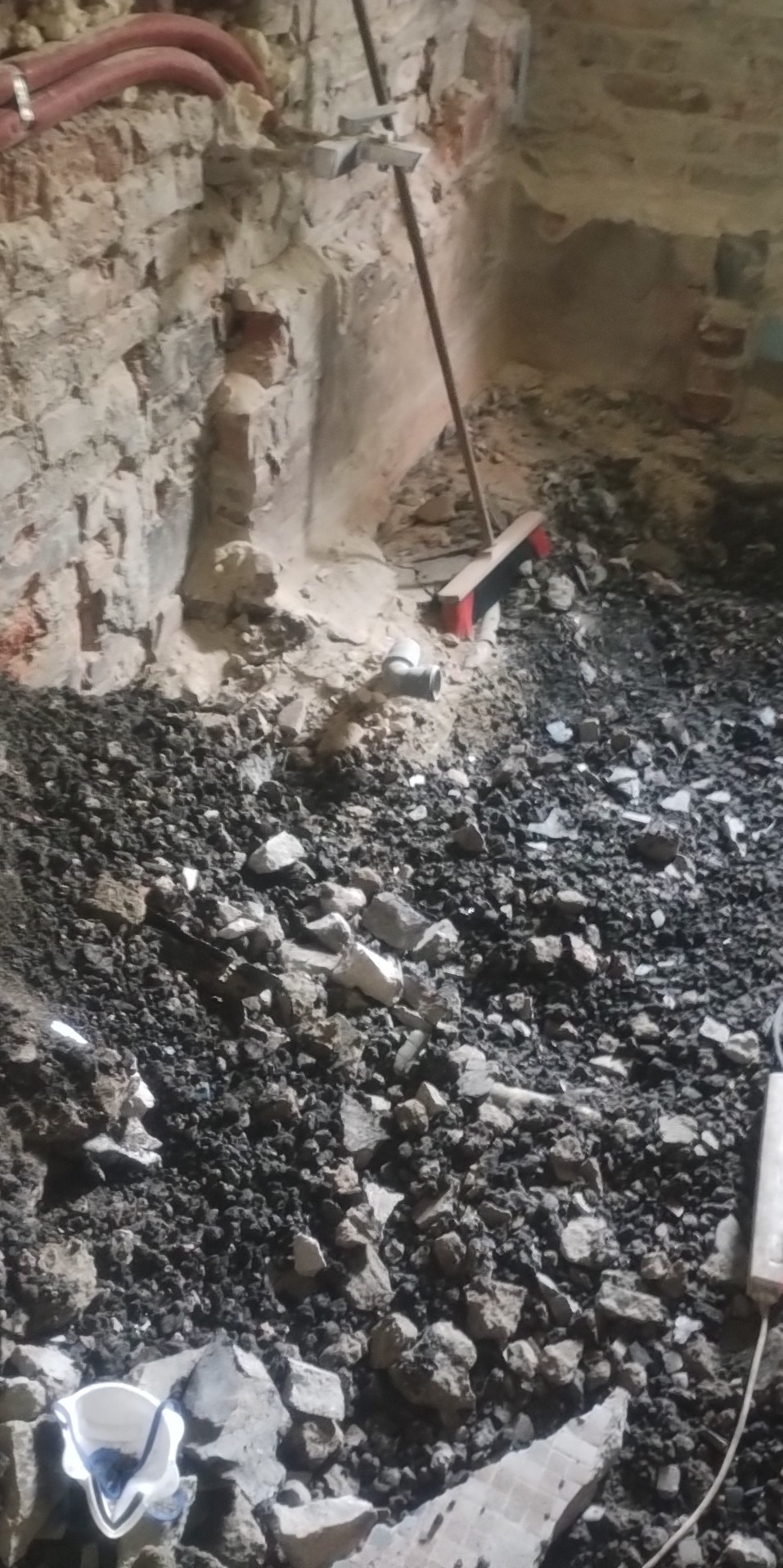 Wnętrze pomieszczenia w trakcie prac rozbiórkowych, widoczne gruzowisko, fragment ściany z cegły i instalacje pod sufitem.