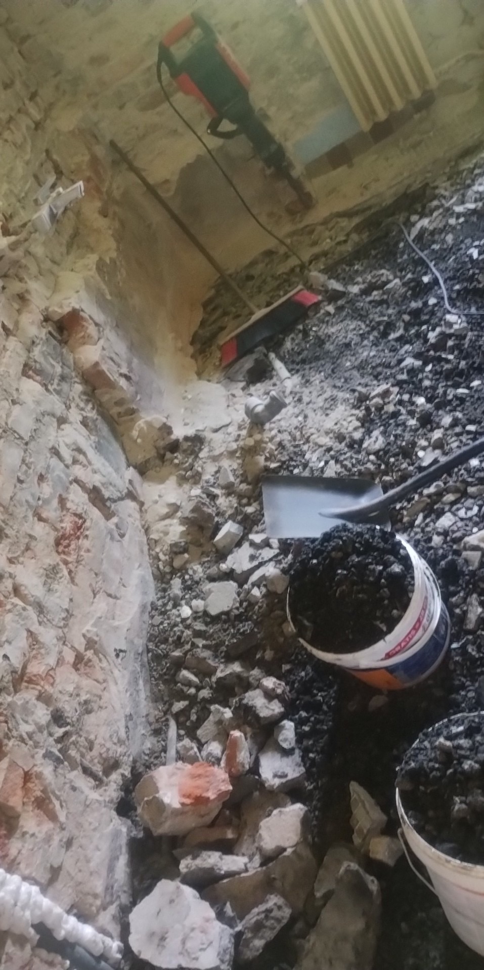 Częściowo wyburzona ściana z cegły, gruz, łopata i kubeł z odpadami, widoczny młot pneumatyczny.