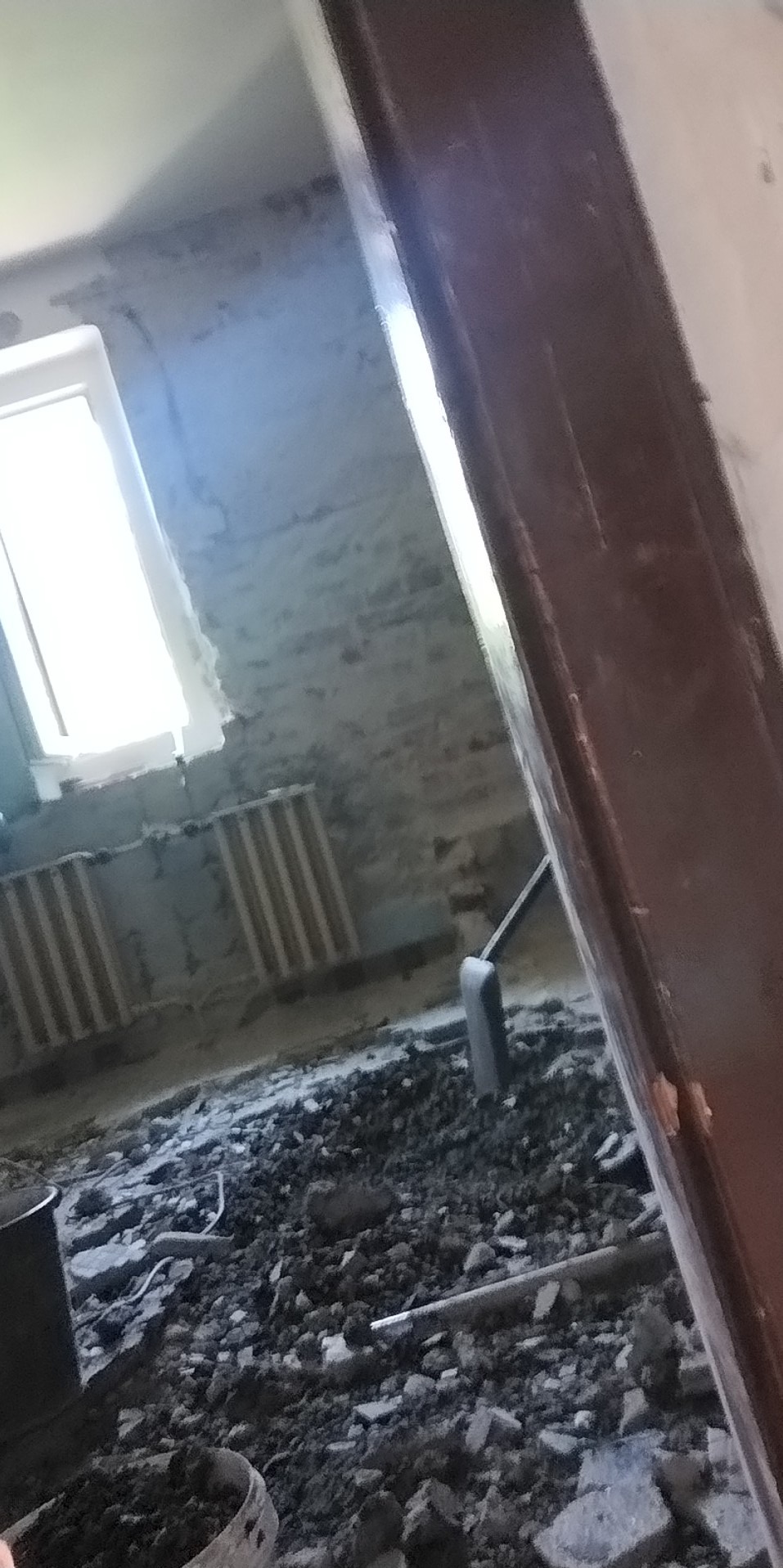 Gruz i odłamki na podłodze w trakcie prac remontowych, widoczny grzejnik i fragment futryny drzwi