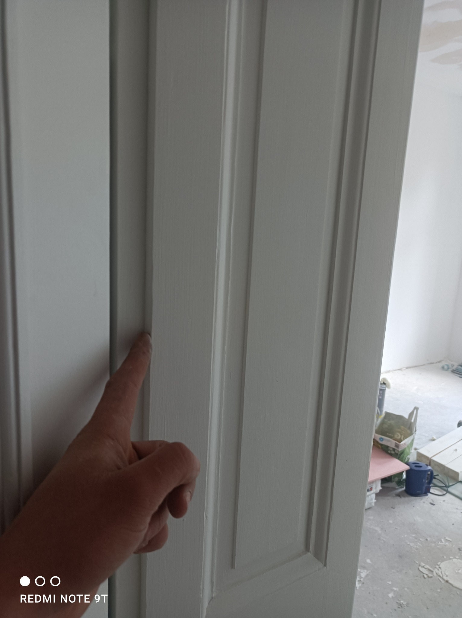 Świeżo pomalowane, białe, profilowane obramowanie drzwi wewnętrznych; palec wskazujący na gładką powierzchnię, w tle widoczne materiały budowlane i resztki gipsu na podłodze.