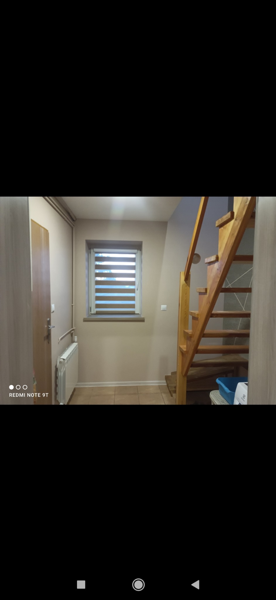 Wnętrze korytarza z drewnianymi schodami, grzejnikiem żeberkowym pod oknem z roletami poziomymi, jasnymi ścianami i brązowymi płytkami na podłodze.