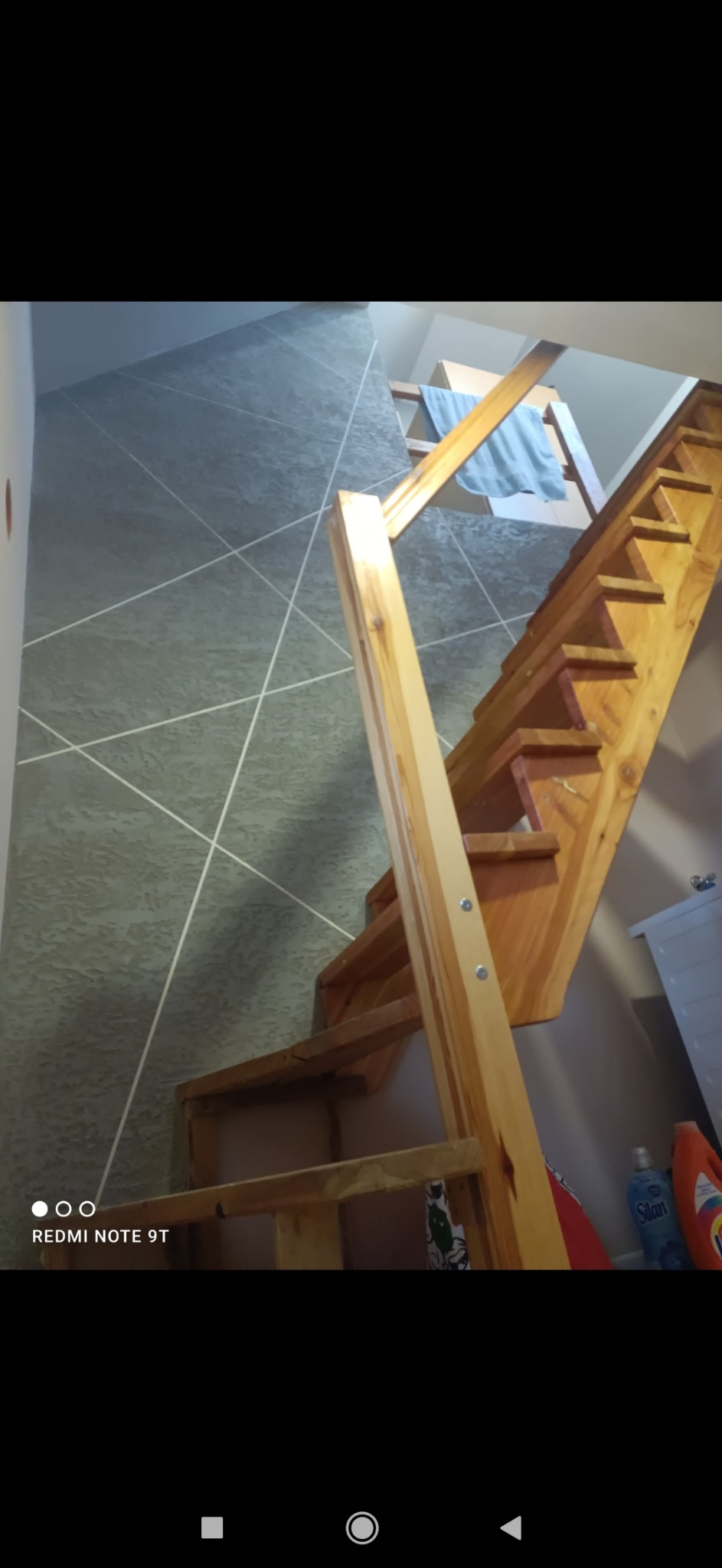 Drewniane schody z surowego drewna, widok z dołu, szare ściany z białymi, ukośnymi liniami dekoracyjnymi w tle.