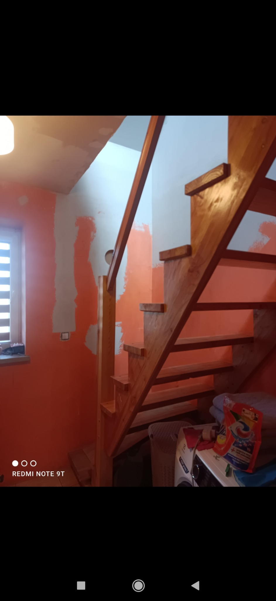 Wnętrze w trakcie odświeżania: drewniane schody na tle pomarańczowej ściany z fragmentami zagruntowanymi na biało, widoczna pralka i proszek do prania