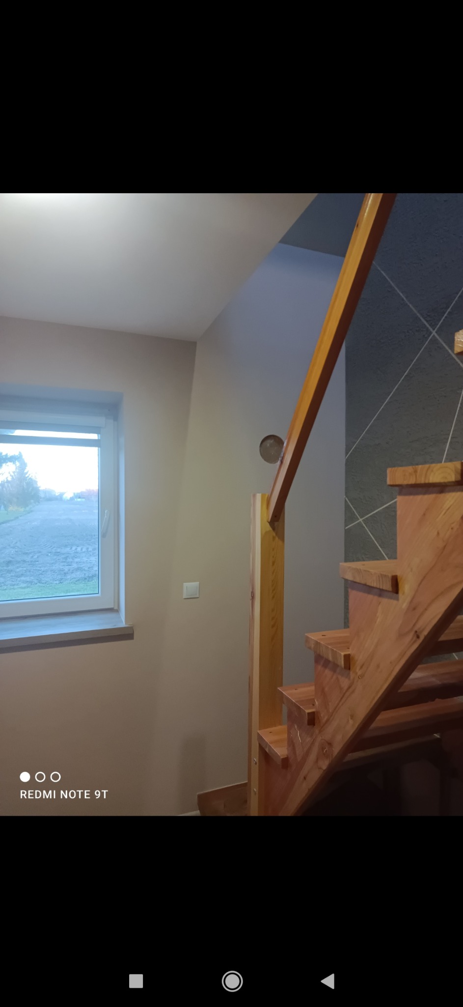 Drewniane schody z balustradą, widoczne z dołu, z fragmentem okna i szarej ściany z geometrycznym wzorem w tle.