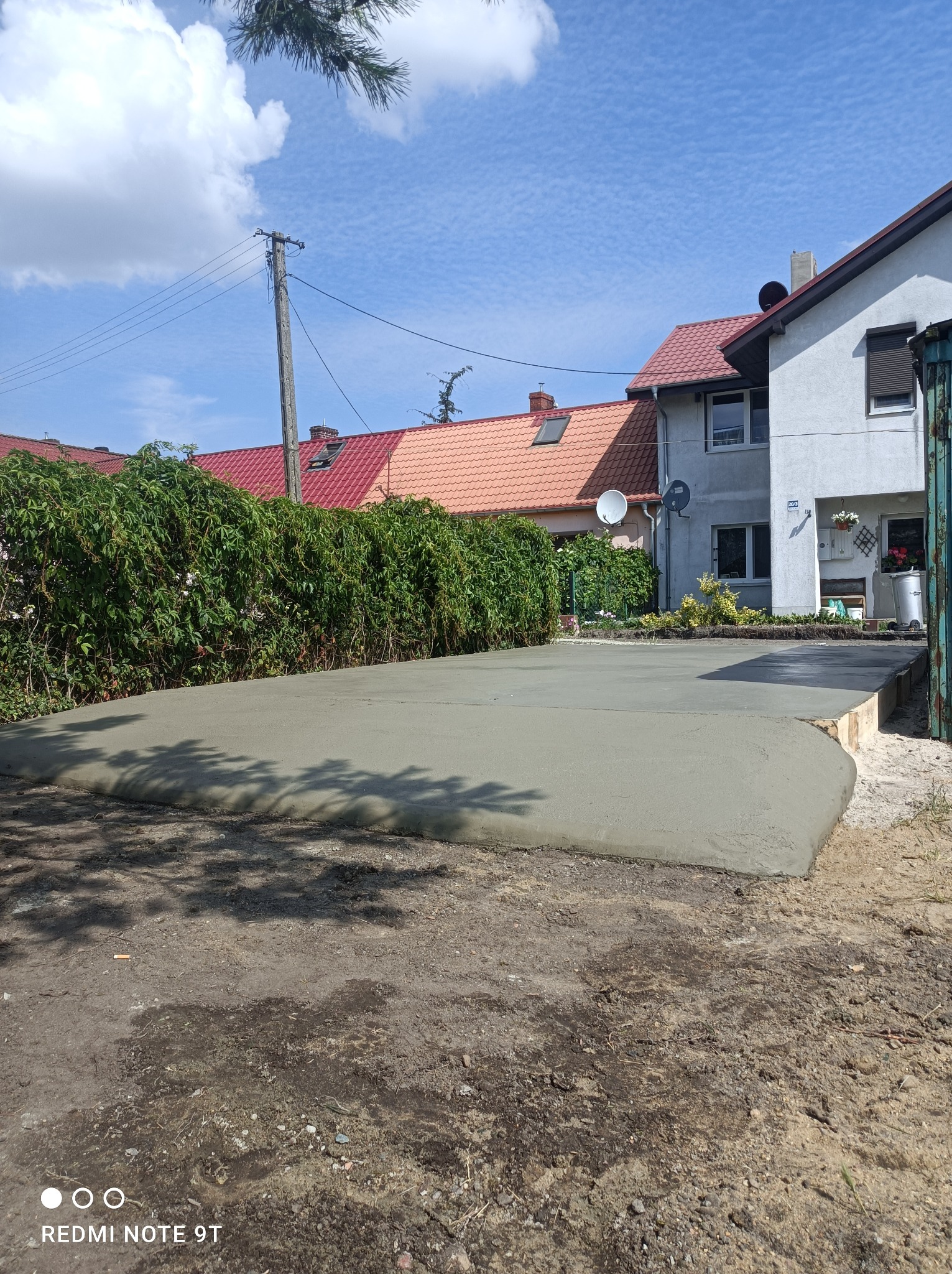 Świeżo wylana, szara betonowa wylewka na podwórku przed domem jednorodzinnym, widoczne drewniane szalunki, w tle zielony żywopłot i błękitne niebo.