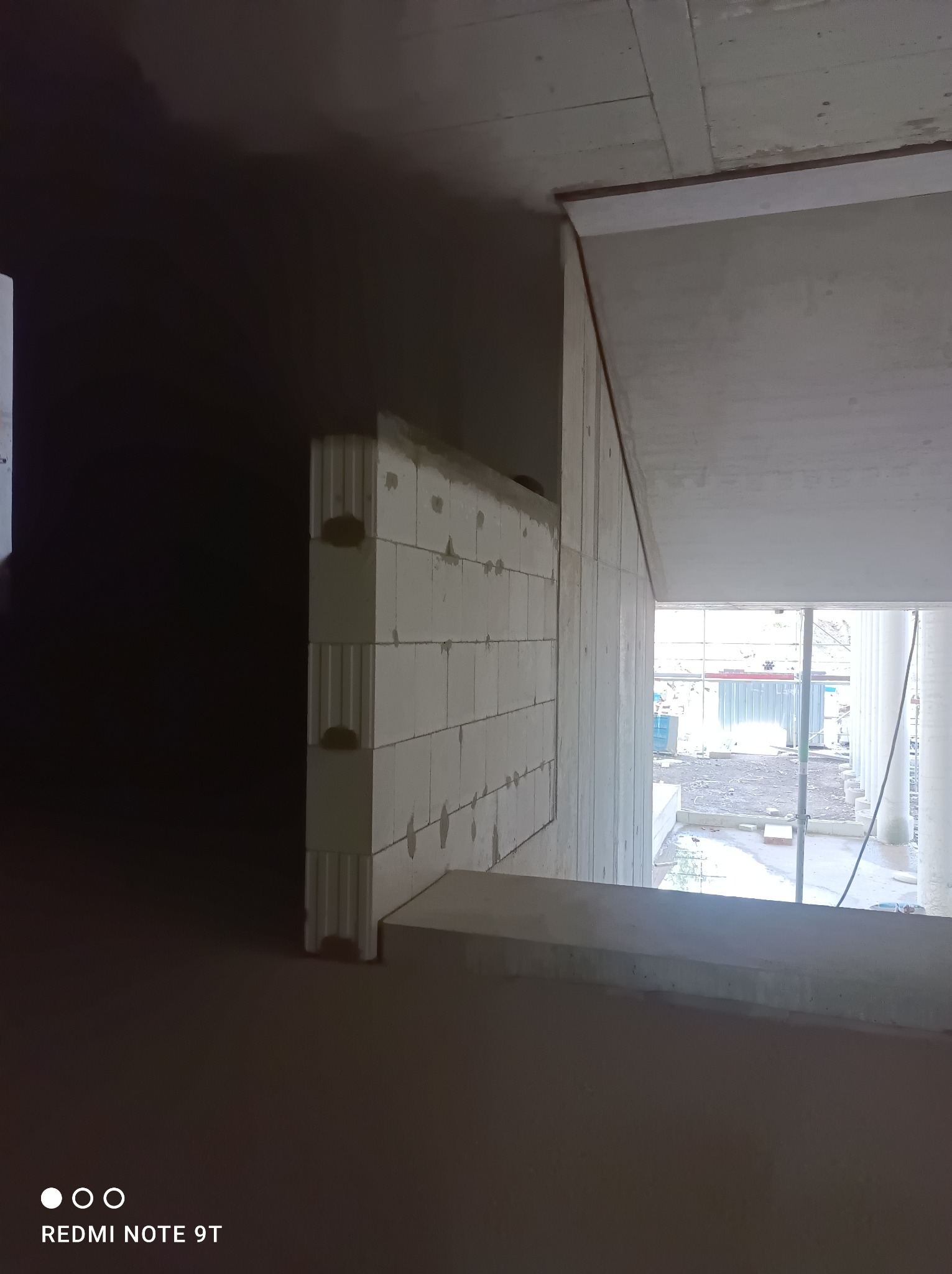 Wnętrze w trakcie budowy: ściana z betonu komórkowego, widoczne łączenia i niedoskonałości, surowy betonowy strop, na zewnątrz rusztowania i materiały budowlane.