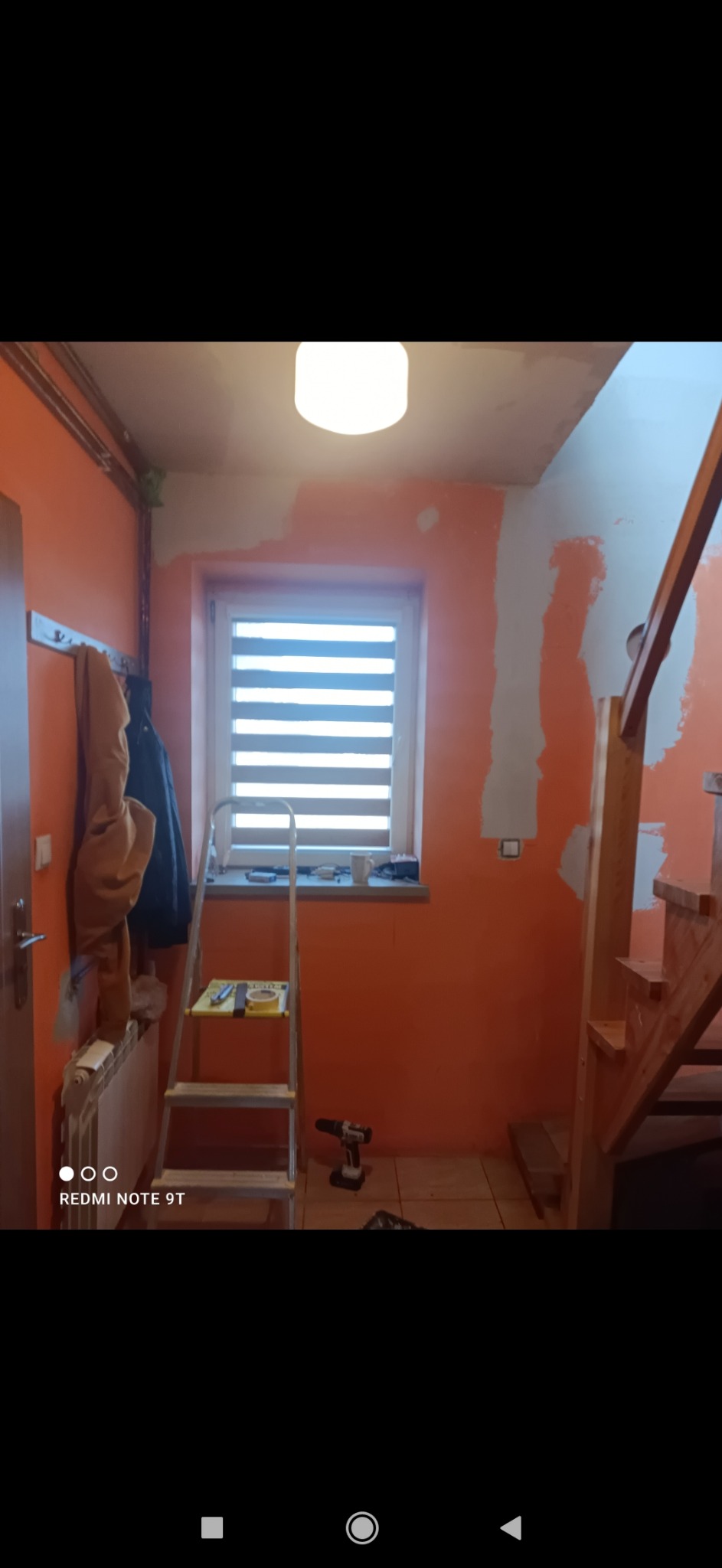 Pomarańczowe wnętrze w trakcie odświeżania: ściany częściowo zagruntowane, drabina z taśmą malarską, wiertarka leżąca na podłodze, widoczne schody drewniane.