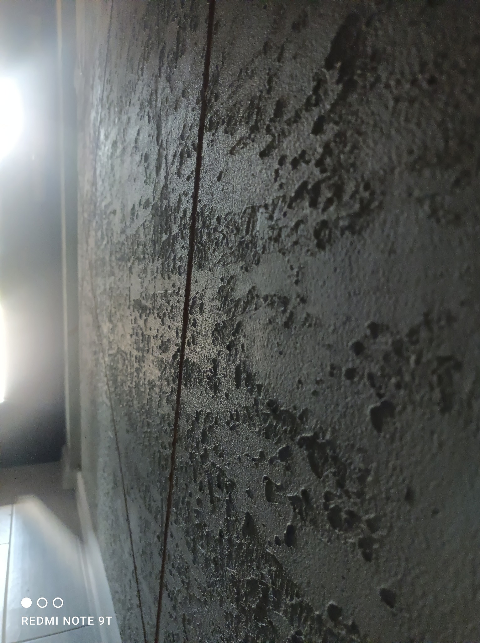 Szara ściana z wyraźną, chropowatą fakturą, oświetlona z lewej strony, z widoczną linią pionową (prawdopodobnie krawędź) i fragmentem podłogi w dolnym rogu.