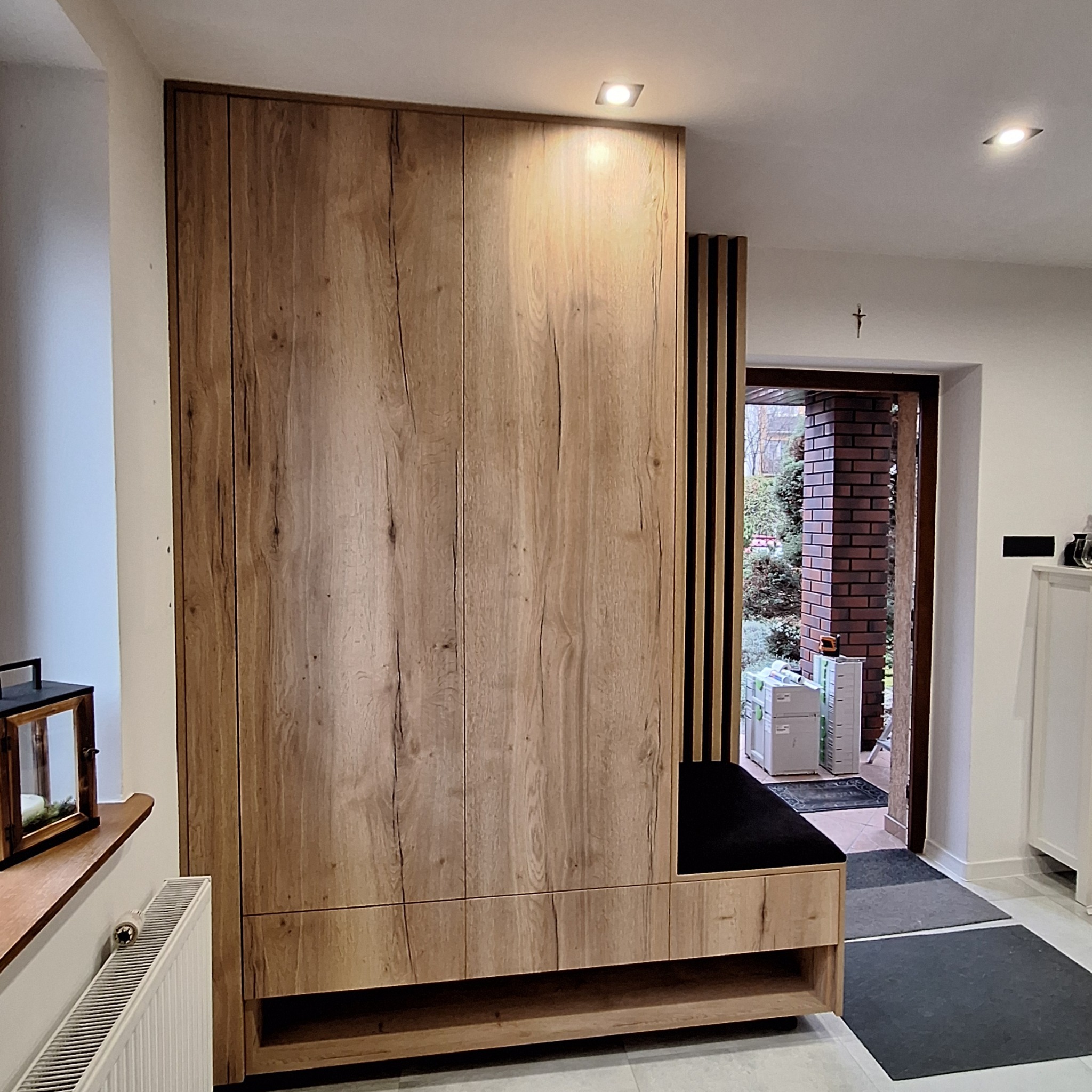 Drewniana szafa z siedziskiem w przedpokoju, widoczne oświetlenie punktowe i dekoracje okienne, zarys wejścia do domu.