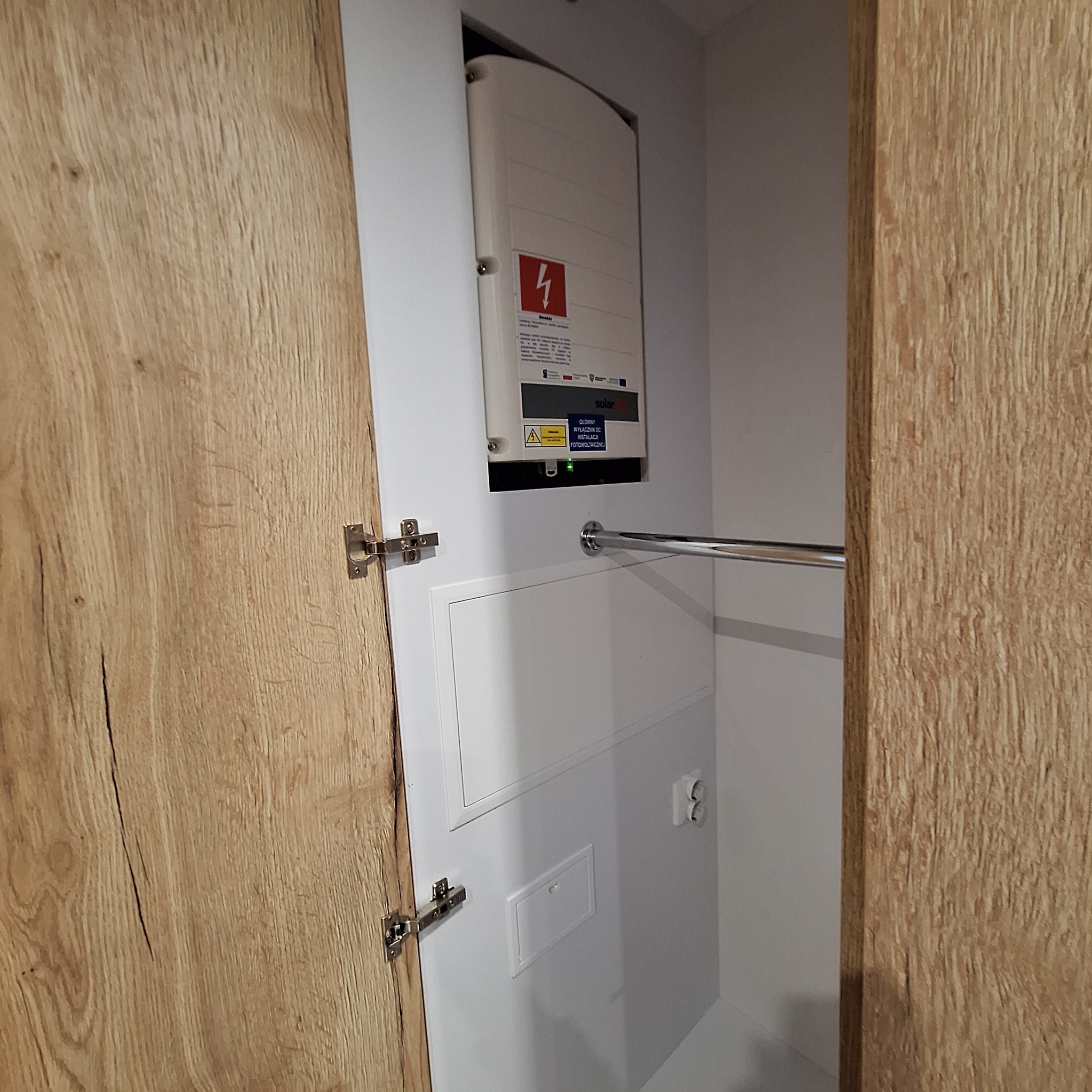 Wnętrze szafy z zamontowanym inwerterem fotowoltaicznym, widoczna drewniana płyta meblowa i metalowy drążek na ubrania.