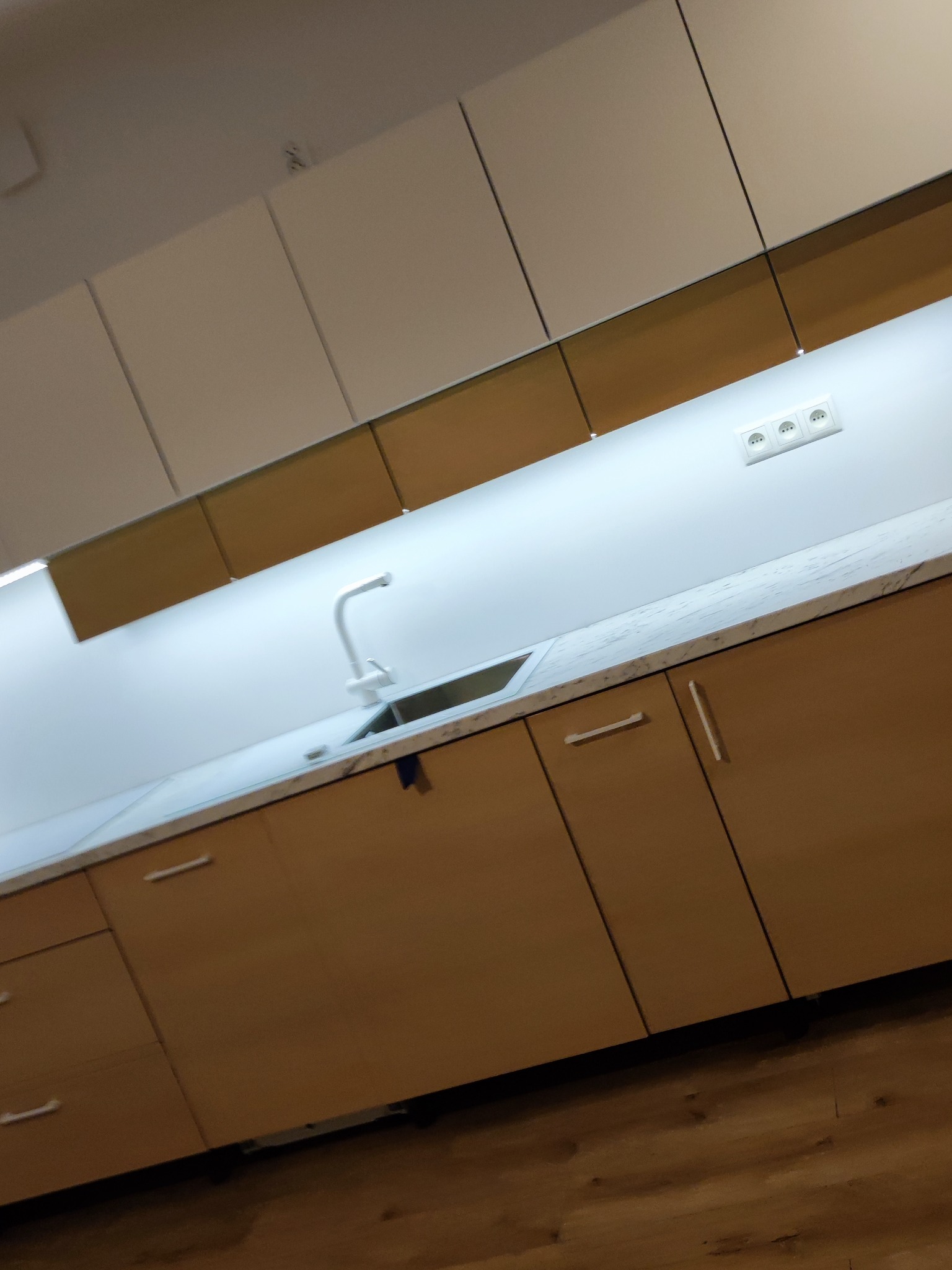 Nowoczesna kuchnia z jasnymi górnymi szafkami i drewnopodobnymi dolnymi, oświetlenie LED pod szafkami, widoczny blat z marmurkowym wzorem i biała bateria przy zlewie.