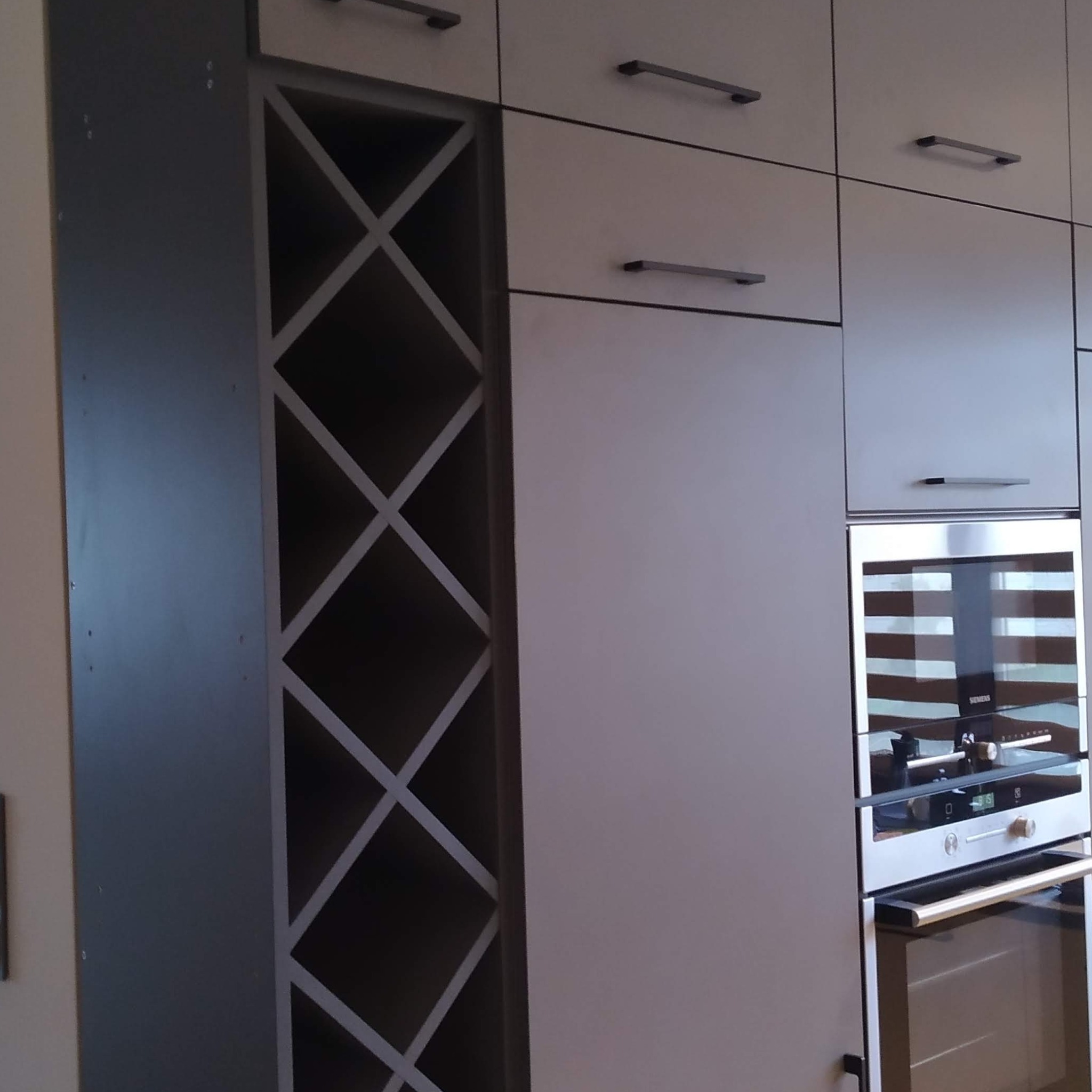 Nowoczesna zabudowa kuchenna z jasnymi szafkami z podłużnymi uchwytami, ciemnoszary regał na wino z geometrycznym wzorem i wbudowany piekarnik Siemens.