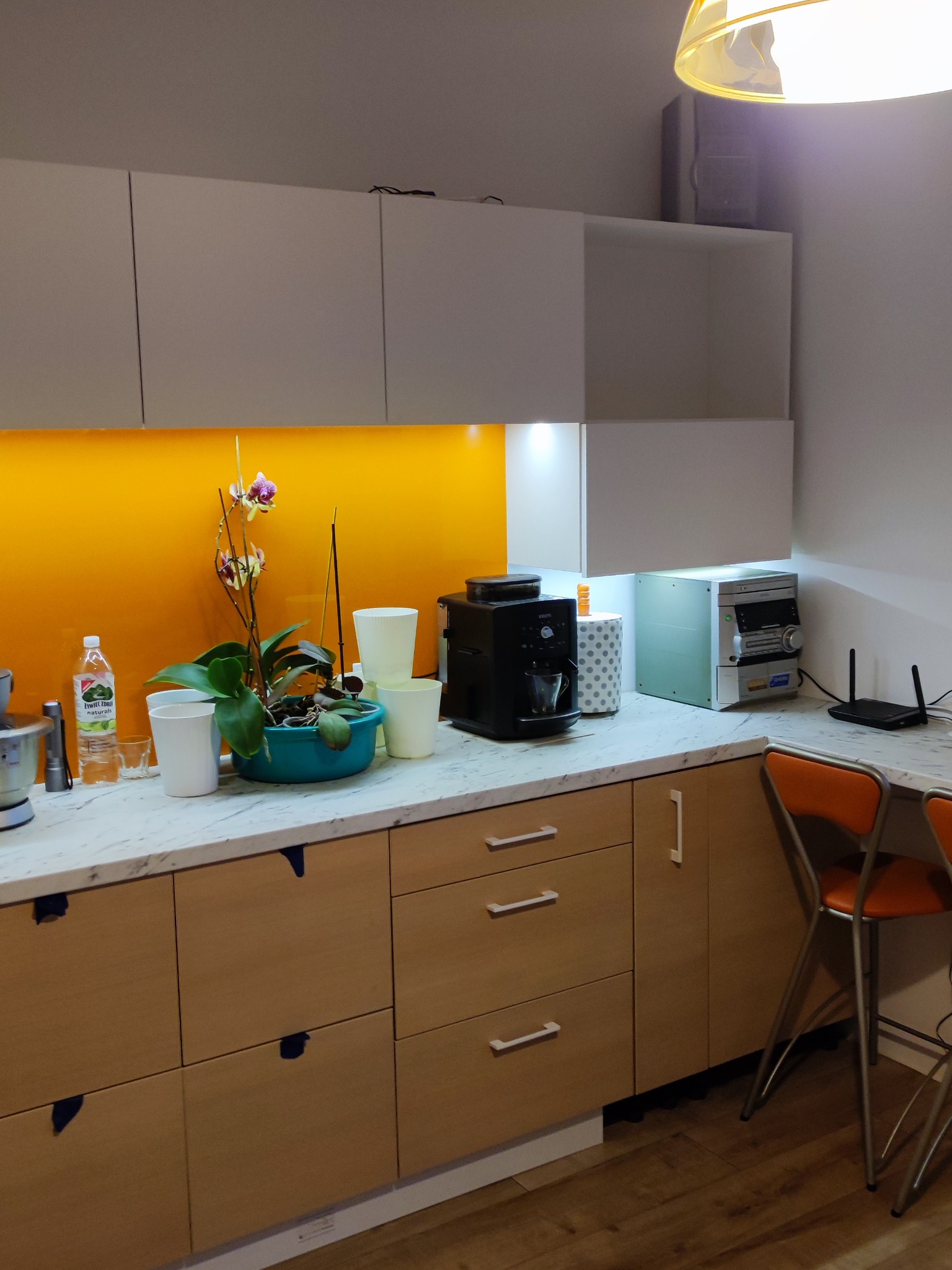 Nowoczesna kuchnia z białymi szafkami górnymi i drewnianymi dolnymi, żółty panel nad blatem, ekspres do kawy, kwiat w doniczce i krzesła barowe.