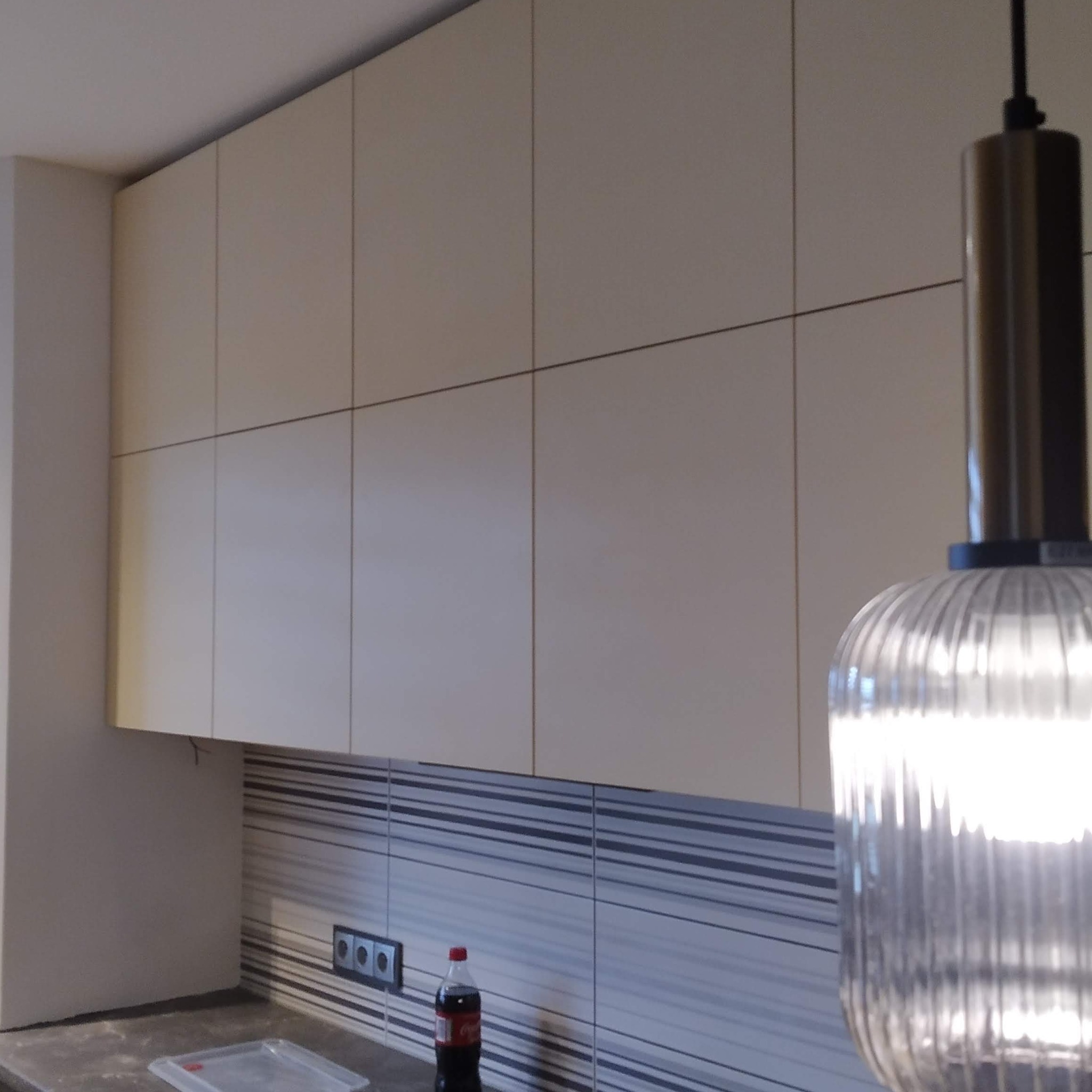 Beżowe górne szafki kuchenne z gładkimi frontami, widoczne gniazdka elektryczne i butelka napoju na blacie, oświetlenie w postaci lampy wiszącej z żarówką.