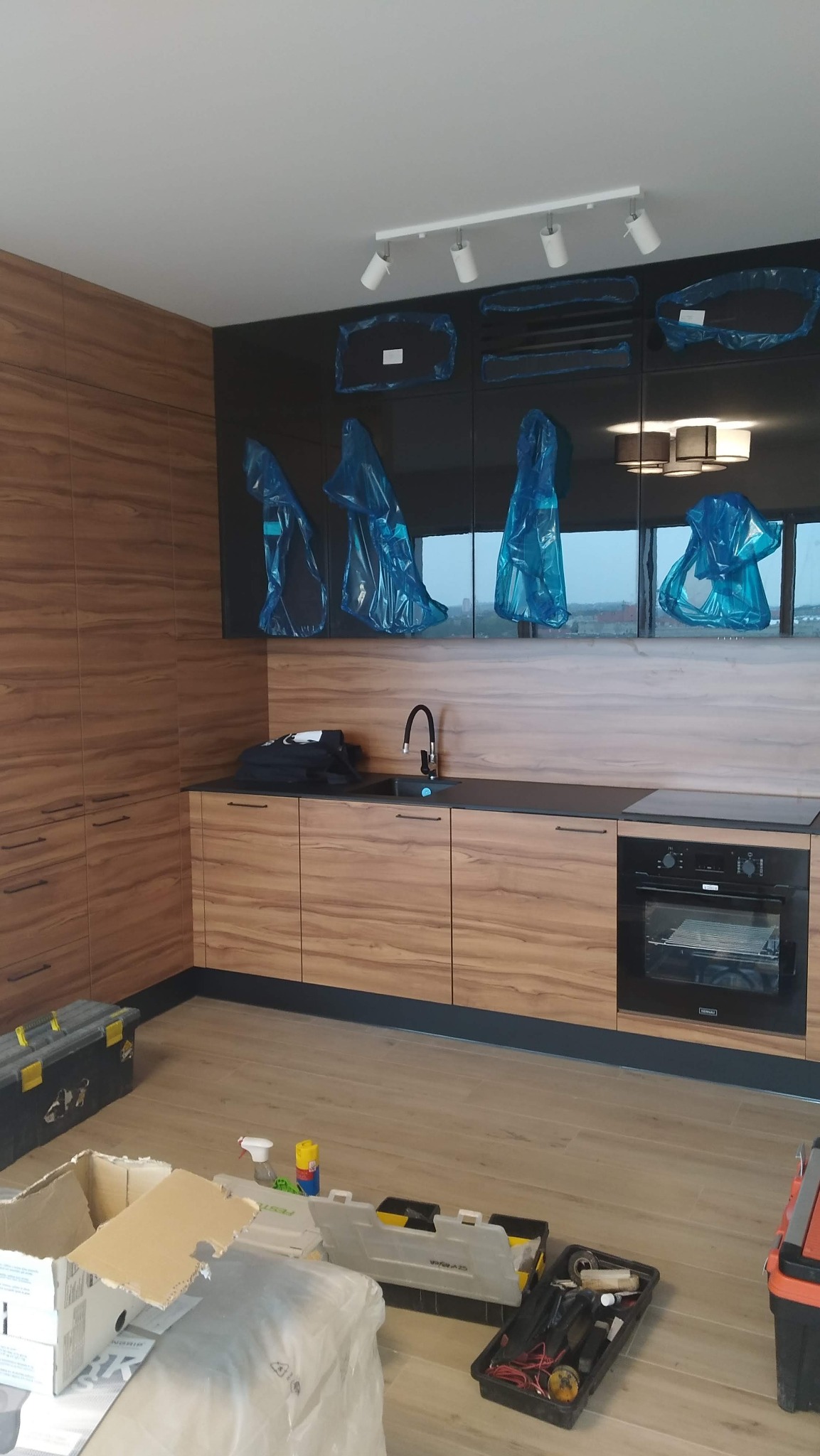 Montaż kuchni: szafki dolne z drewnopodobnym frontem, czarny blat i piekarnik, szafki wiszące z czarnymi, błyszczącymi frontami zabezpieczonymi folią, widoczne narzędzia pracy.