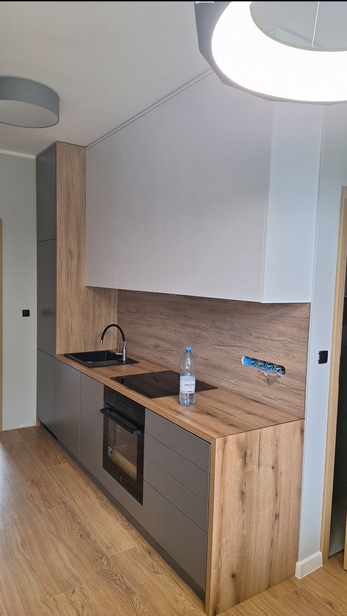 Nowoczesna kuchnia z szarymi szafkami dolnymi, drewnopodobnym blatem i białymi szafkami górnymi, z widocznym zlewem, płytą grzewczą i butelką wody mineralnej na blacie.