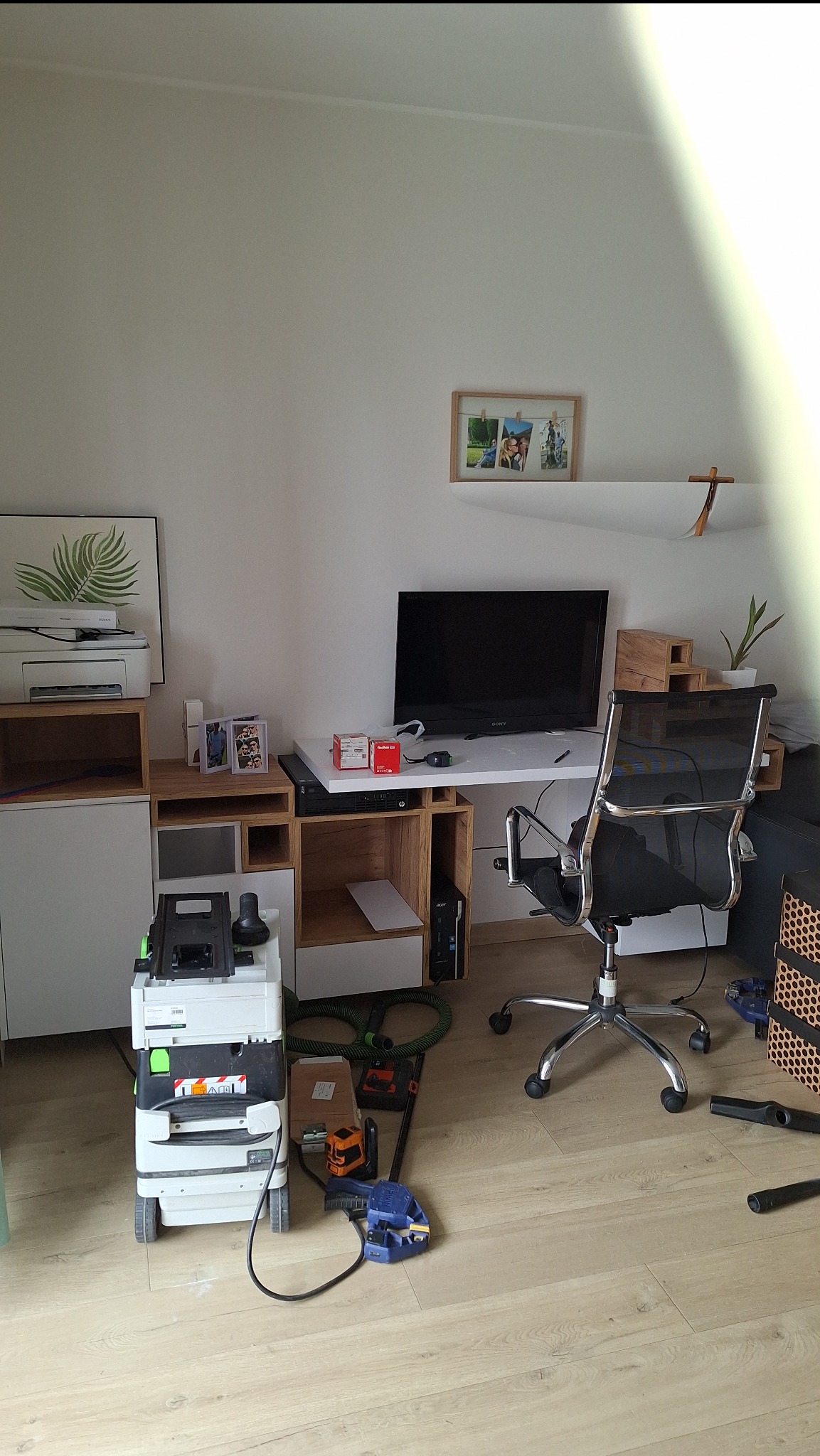 Fragment pokoju z biurkiem, krzesłem biurowym i regałem z półkami, na podłodze leżą narzędzia i odkurzacz przemysłowy.