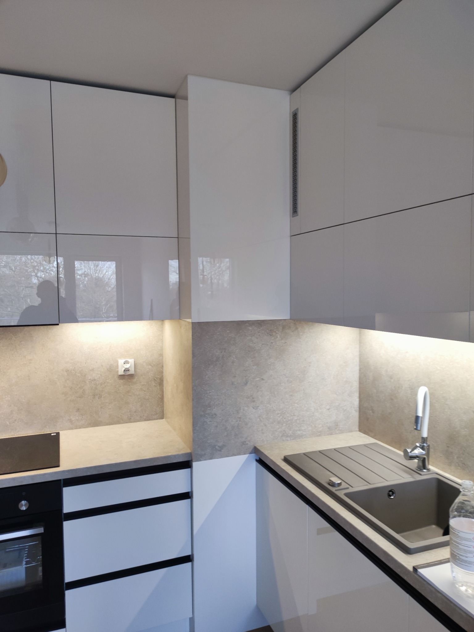 Nowoczesna kuchnia z białymi, lakierowanymi frontami szafek, szarym blatem i zlewozmywakiem, oświetleniem podszafkowym i widocznym odbiciem okna w górnych szafkach.